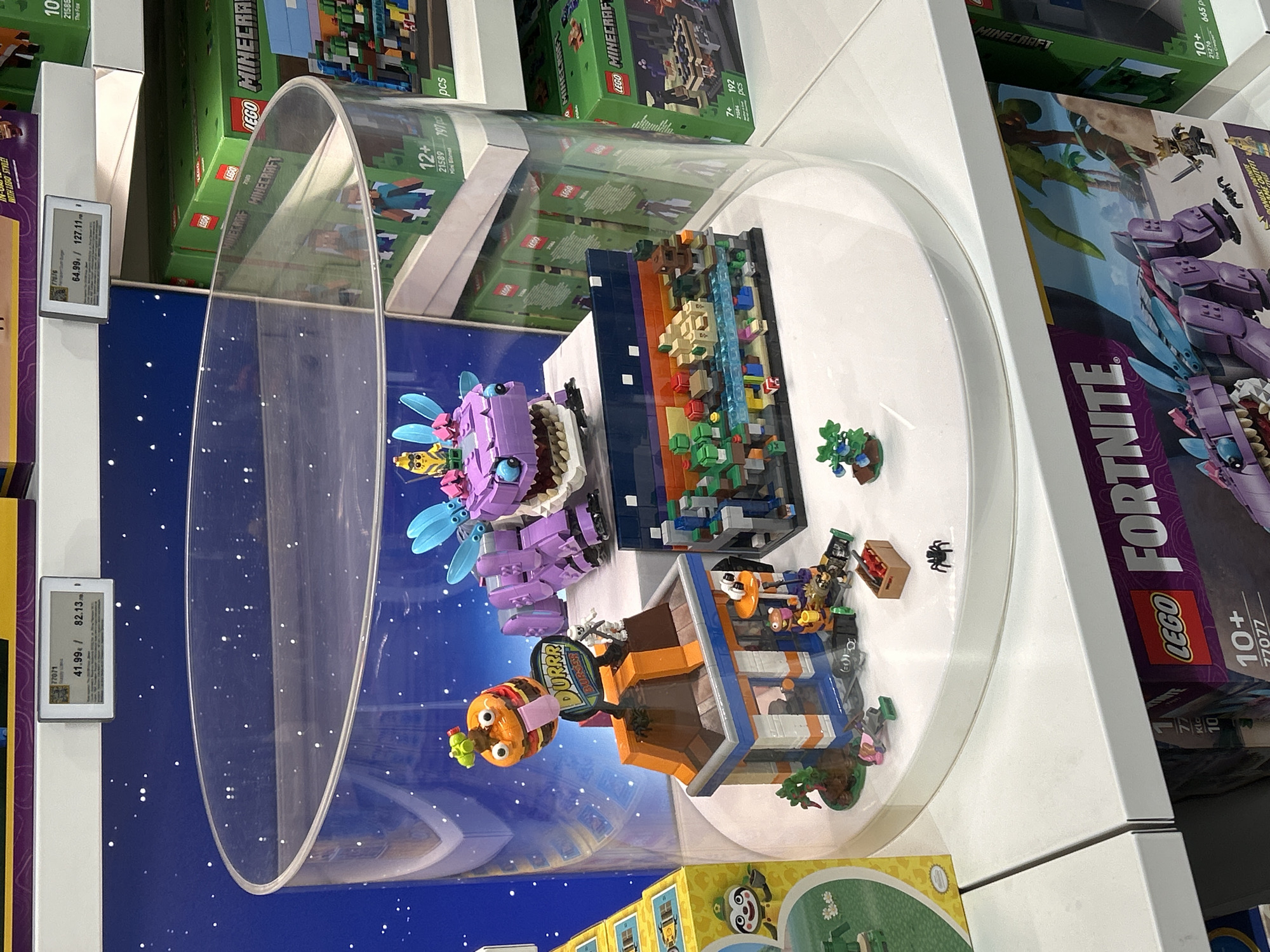 The image shows a display of LEGO sets inside a store, likely a toy store or department store. The display is set within a clear acrylic dome on a white shelf. Inside the dome are assembled LEGO sets, including a Fortnite set and a Minecraft set.
To the left of the display, price tags are visible, indicating product codes and prices in Euros. The Fortnite set packaging is visible to the right, showing the LEGO logo and a "10+" age recommendation.
Other LEGO Minecraft sets are stacked above the dome. The environment appears to be a retail space with bright lighting. The location is not discernible.