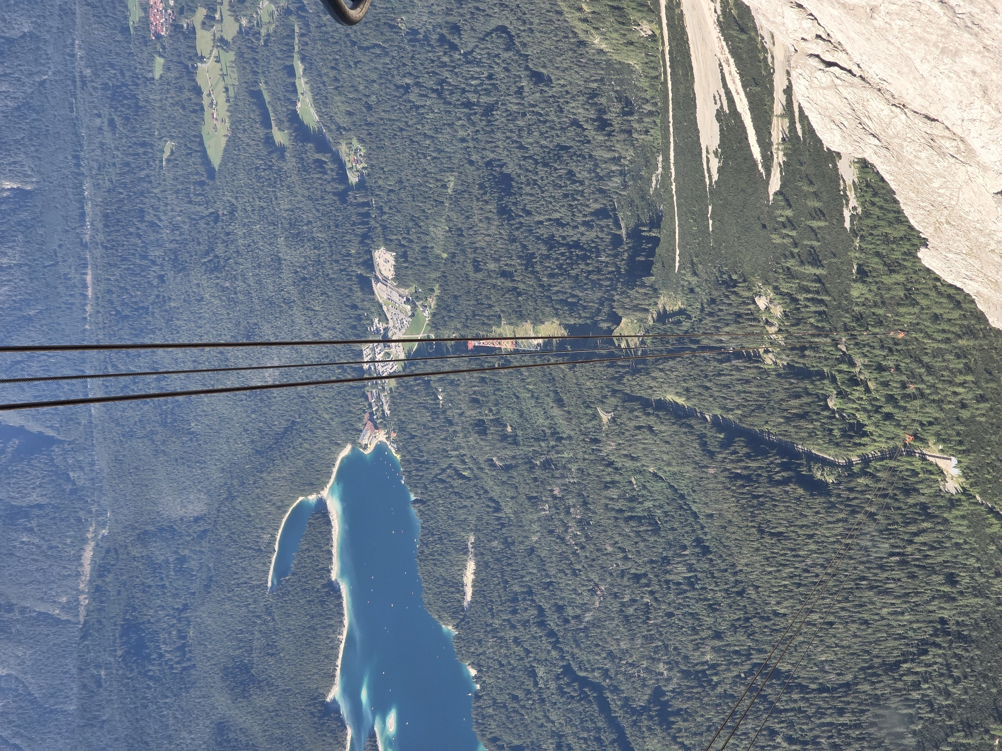 The image captures a scenic aerial view of the Zugspitze region near Garmisch-Partenkirchen, Germany. In the foreground, rough, rocky terrain dominates the right side, contrasting with the lush green coniferous forest that covers the slopes below. A body of clear blue water, likely the Eibsee, is visible in the upper left quadrant, with a small island and several boats dotting its surface. A cluster of buildings, possibly a village or resort, is situated on the edge of the lake. Several thick cables, likely part of a cable car system, descend vertically through the frame, indicating a popular tourist area. In the middle ground, a red and white pylon for the cable car is visible among the trees, with a road winding through the forest. The overall scene depicts a vast, mountainous landscape characterized by dense forests, a pristine lake, and infrastructure for tourism.