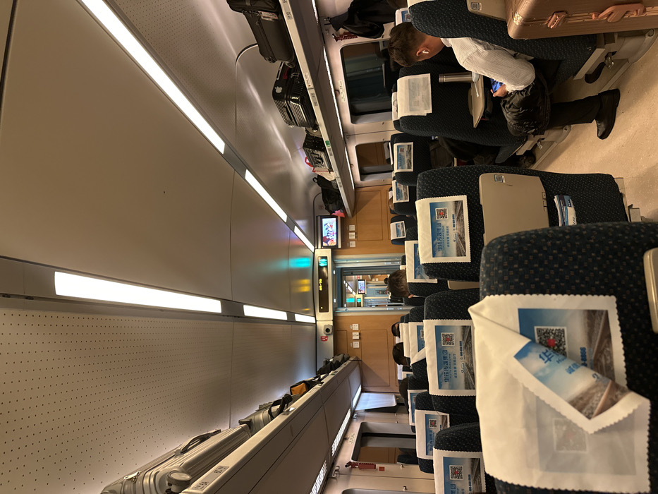 The scene depicts the interior of a modern passenger train car, believed to be in or near Tonglu County, China. The perspective is from a seat in the mid-section of the car, looking forward towards the end of the carriage.
The train interior features rows of dark blue fabric seats with a subtle patterned design, each fitted with a white, disposable headrest cover. Many headrest covers display a Chinese advertisement from "China Railway Media Group" featuring train tracks, a QR code, and a phone number (400-886-8888). Overhead luggage racks line both sides of the car, filled with various suitcases, bags, and backpacks, including a prominent silver metallic suitcase on the left rack and a black case with an Apple logo sticker on the right rack.
The ceiling is light-colored, featuring two prominent linear LED light fixtures running the length of the car, providing bright illumination. A perforated ceiling section is visible on the left. The floor is light-colored.
On the right side of the frame, a male passenger is seated facing away from the camera. He wears a light-colored, long-sleeved shirt and dark trousers, with black shoes. He has white wireless earbuds in his ears and is actively using a mobile phone held in both hands. A small, light-colored tray table is extended from the seat in front of him, holding a metallic water bottle. A brown hard-shell suitcase rests on the floor beside his seat.
In the distance, at the front of the car, a doorway leads to an adjacent carriage or vestibule. Above this doorway, a digital display shows "08:56" alongside green and red indicator lights. A smaller screen displaying visual content is mounted on the wall to the right of the doorway. A red fire extinguisher is visible mounted on the wall to the left of the doorway. Several other passengers are partially visible further down the aisle, seated and largely obscured by the headrests.