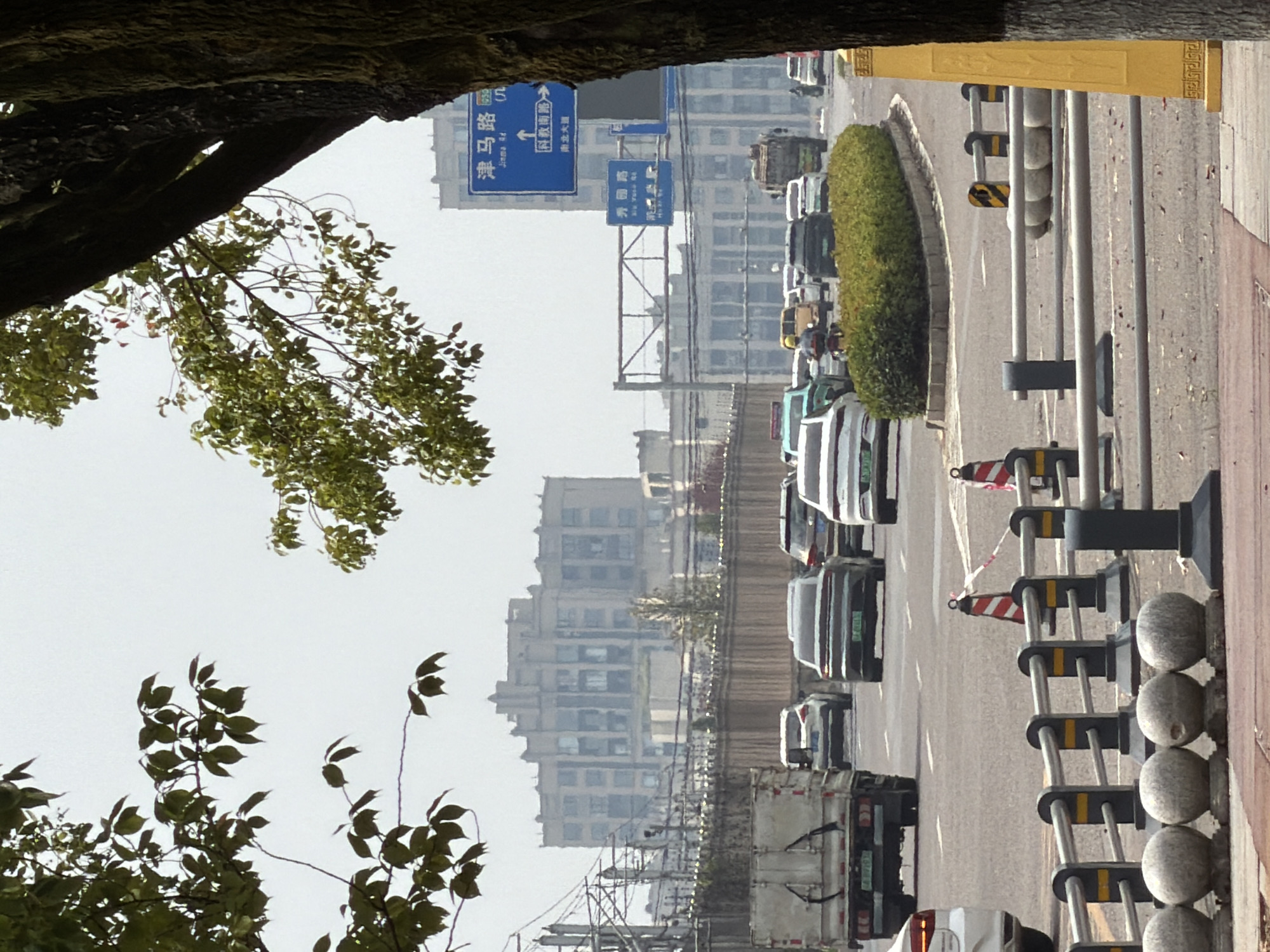 The image depicts a street scene in Jiangjin District, Chongqing, China, likely on a hazy day. In the foreground, a paved sidewalk with decorative stone spheres is visible, bordered by metal railings and construction barriers. The street itself features multiple lanes of traffic with various vehicles, including cars, a truck, and a motorcycle.
A prominent blue road sign with Chinese characters and arrows is positioned above the street, indicating directions to "Jinma Rd" and "Nan'an Ave." Another smaller blue sign displays "Bai'an Rd." In the background, a large, multi-story building with an imposing architectural style dominates the skyline. Trees with green foliage frame the scene from the left and right, partially obscuring the sky and buildings. The overall atmosphere suggests a typical urban environment during daylight hours.