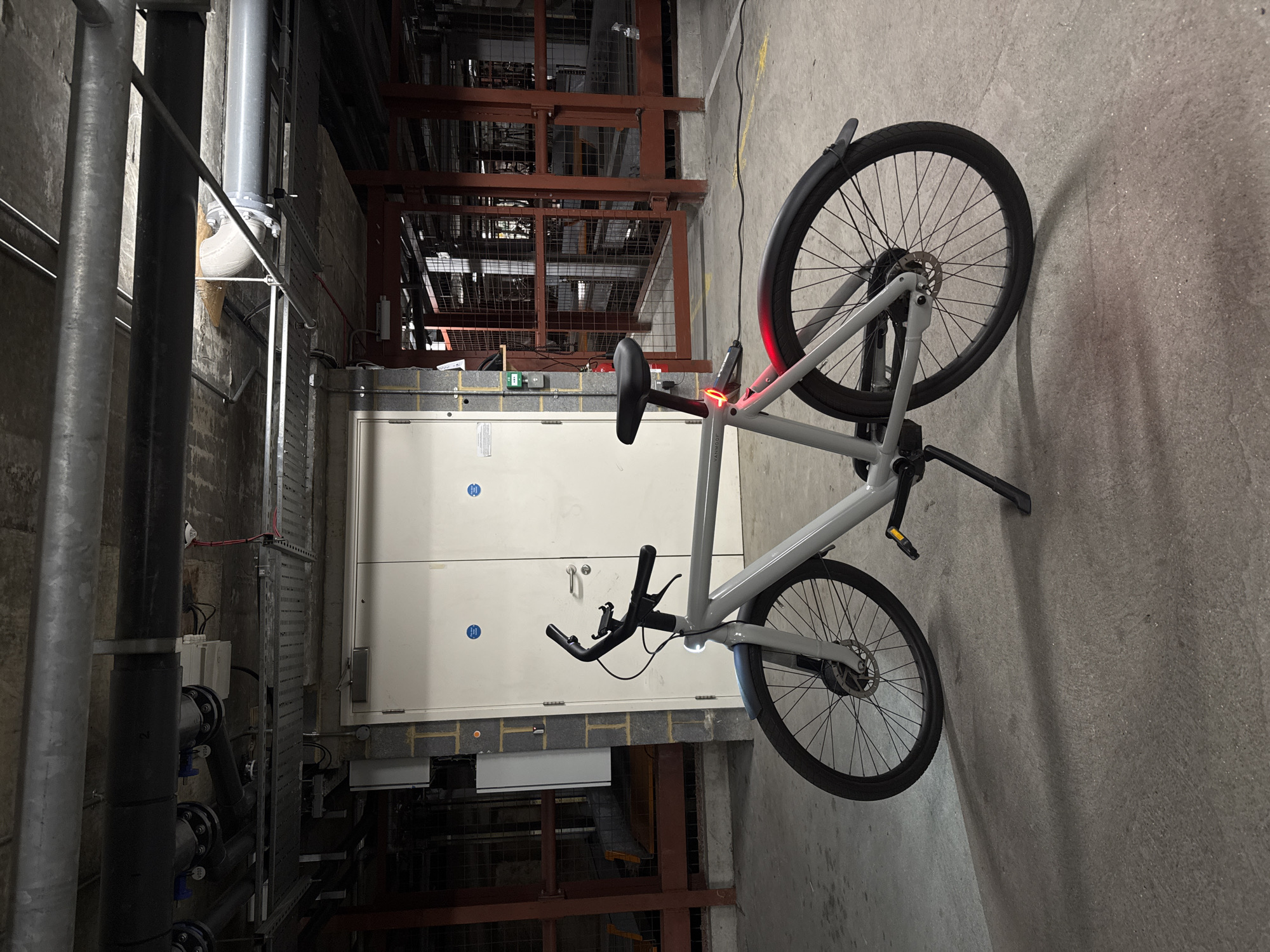 A grey VanMoof electric bicycle is prominently displayed in an indoor, industrial-style setting. The bike is positioned on a grey concrete floor, seen from its left side, with a black charging cable connected to its frame near the rear wheel. Its integrated rear light is illuminated red, indicating it is currently charging. The bicycle features a minimalist design, disc brakes, and black components like the saddle, handlebars, and mudguards. The brand name "VanMoof" is faintly visible on the frame.
The background reveals a functional utility area. To the left of the bike, a complex network of large grey pipes, both horizontal and vertical, runs alongside a textured wall, likely made of brick or concrete. Further back, a large, light-colored double-doored cabinet or electrical panel is visible, adorned with two blue circular warning or informational stickers. Below this cabinet, there's an opening revealing more industrial equipment.
On the right side of the background, a dark, rusty-red metal framework or shelving structure fills the upper portion of the image. Faint yellow markings are visible on the concrete floor in the upper right quadrant. The lighting is dim, suggesting an artificial light source within a service or maintenance area, possibly in a basement or utility room. The overall impression is one of a practical, non-decorative space, typical of building infrastructure in an urban environment like London, United Kingdom.