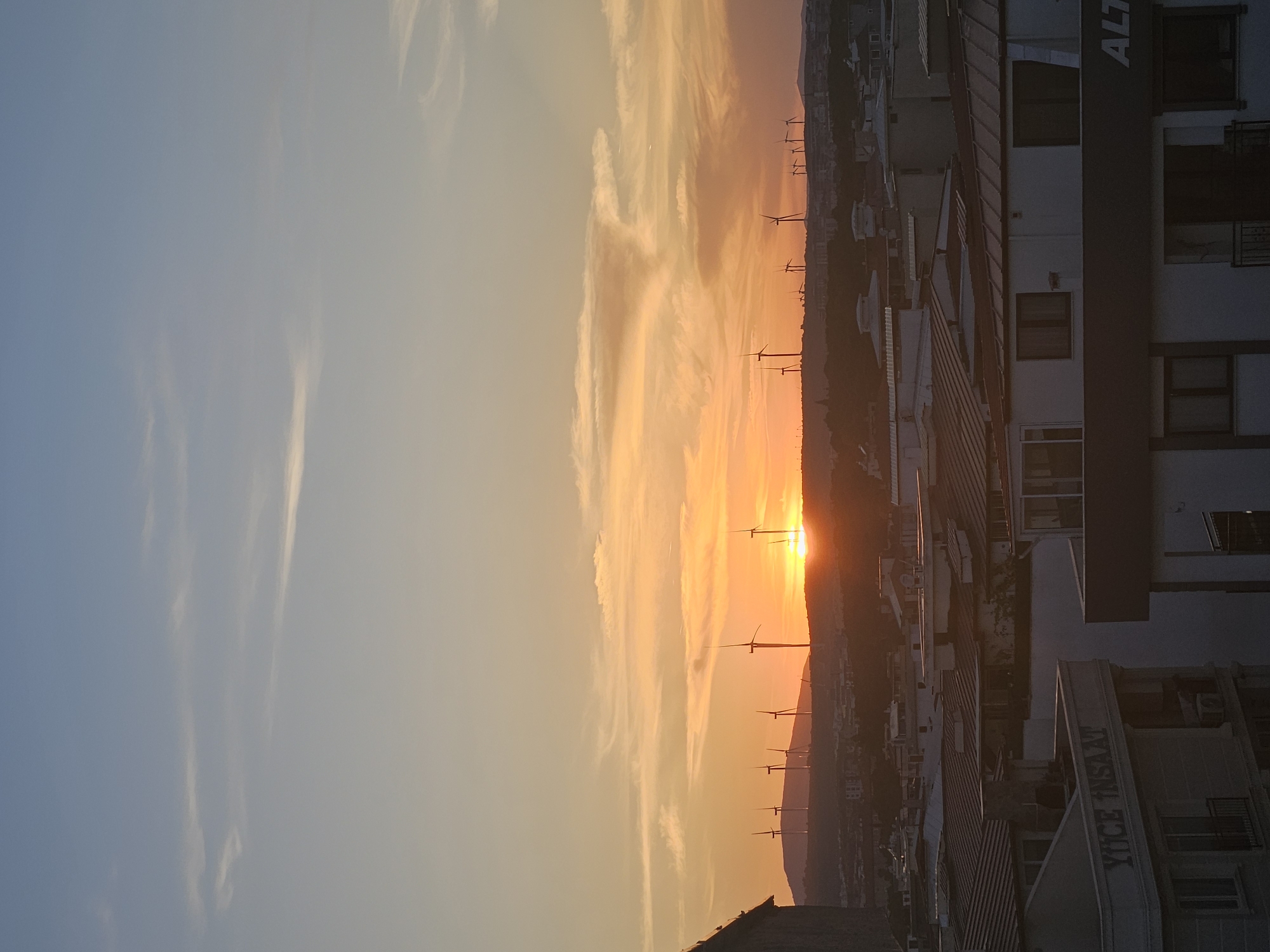 The image depicts a sunset over the city of Seferihisar, Turkey. In the foreground, several buildings with tiled roofs are visible. One building on the left has text that reads "YÜCE İNŞAAT". Another building on the right has the partial text "ALT". In the middle ground, a collection of buildings and structures form the urban landscape. The background is dominated by a hill with numerous wind turbines silhouetted against the colorful sunset sky. The sun is low on the horizon, casting a warm orange glow. The sky features streaks of orange, yellow, and blue, with some wispy clouds. There are no people visible in the image.