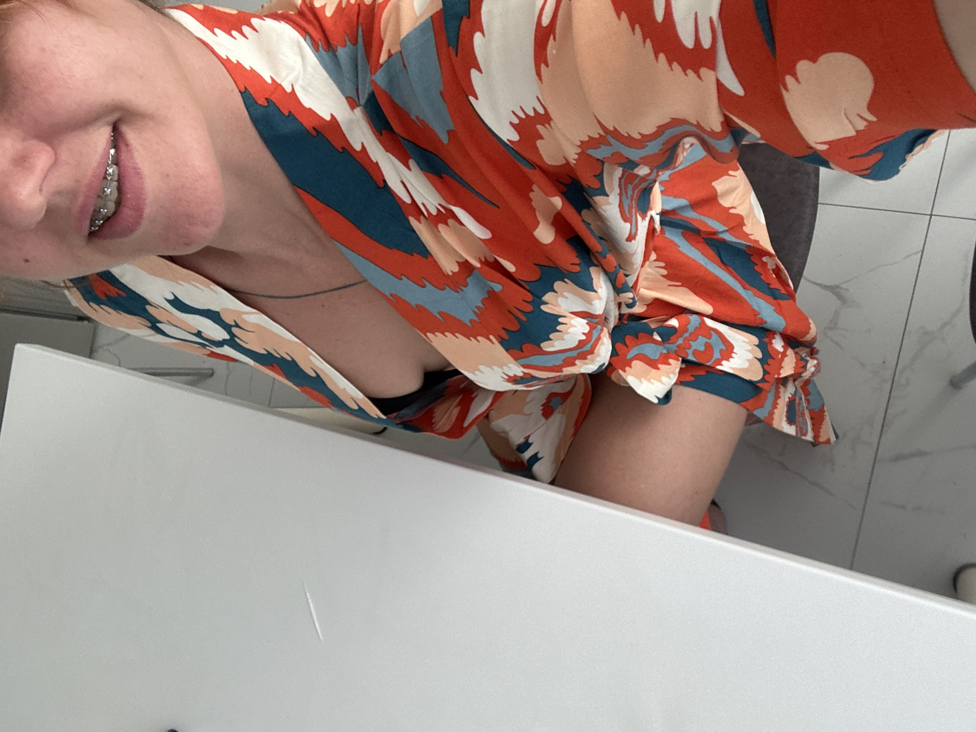 A woman with braces smiles in a brightly lit room. She wears a colorful, patterned wrap dress with orange, blue, white, and peach elements. The woman is indoors, possibly in a bathroom, as a white counter or sink is prominent in the foreground, and white tiles are visible in the background. The location is not evident from the image.