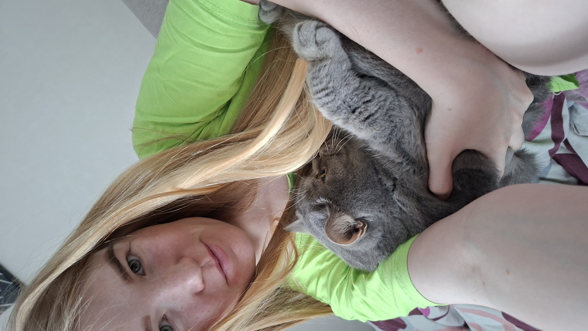 A woman with long blonde hair and wearing a neon green shirt is holding a gray cat. The cat is nestled in her arms. Her face is visible, showing green eyes. The background is out of focus and appears to be a bedroom with bedding. The location is Chaikovsky, Russia.