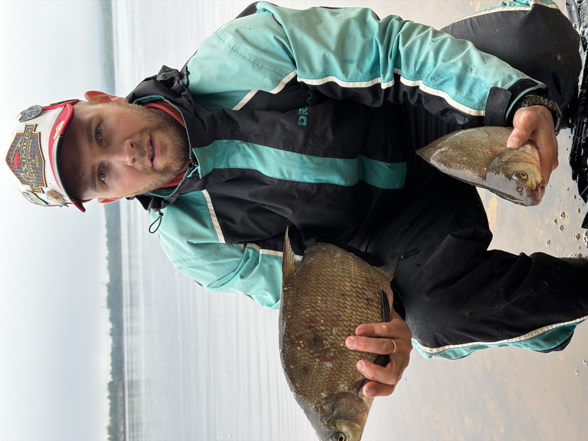 A man is depicted outdoors, crouching or kneeling by the edge of a large, calm body of water, likely a lake or wide river, in Chaikovsky, Russia. He holds two flat-bodied fish, one in each hand, suggesting he has just caught them while fishing. The man has a beard and is looking directly at the camera with a neutral expression.
He is wearing a black and teal outdoor jacket with white stripes, and dark pants. A dark-colored watch is visible on his left wrist, and a simple gold wedding band is on his right ring finger. He wears a baseball cap that is white, red, and dark-colored, featuring several pins. Text on the cap reads "MAXIMUS" and "THE BEST SPINNING & FLY RODS." Part of the word "DRAGON" is visible on the chest area of his jacket.
The two fish appear to be bream, with the one in his left hand being noticeably larger than the one in his right. The water surface is still, reflecting the soft, muted light of what appears to be an overcast daytime. A faint, distant tree line is visible across the water, indicating a natural, expansive outdoor setting. The overall weather seems damp or cool, consistent with the man's protective outerwear.