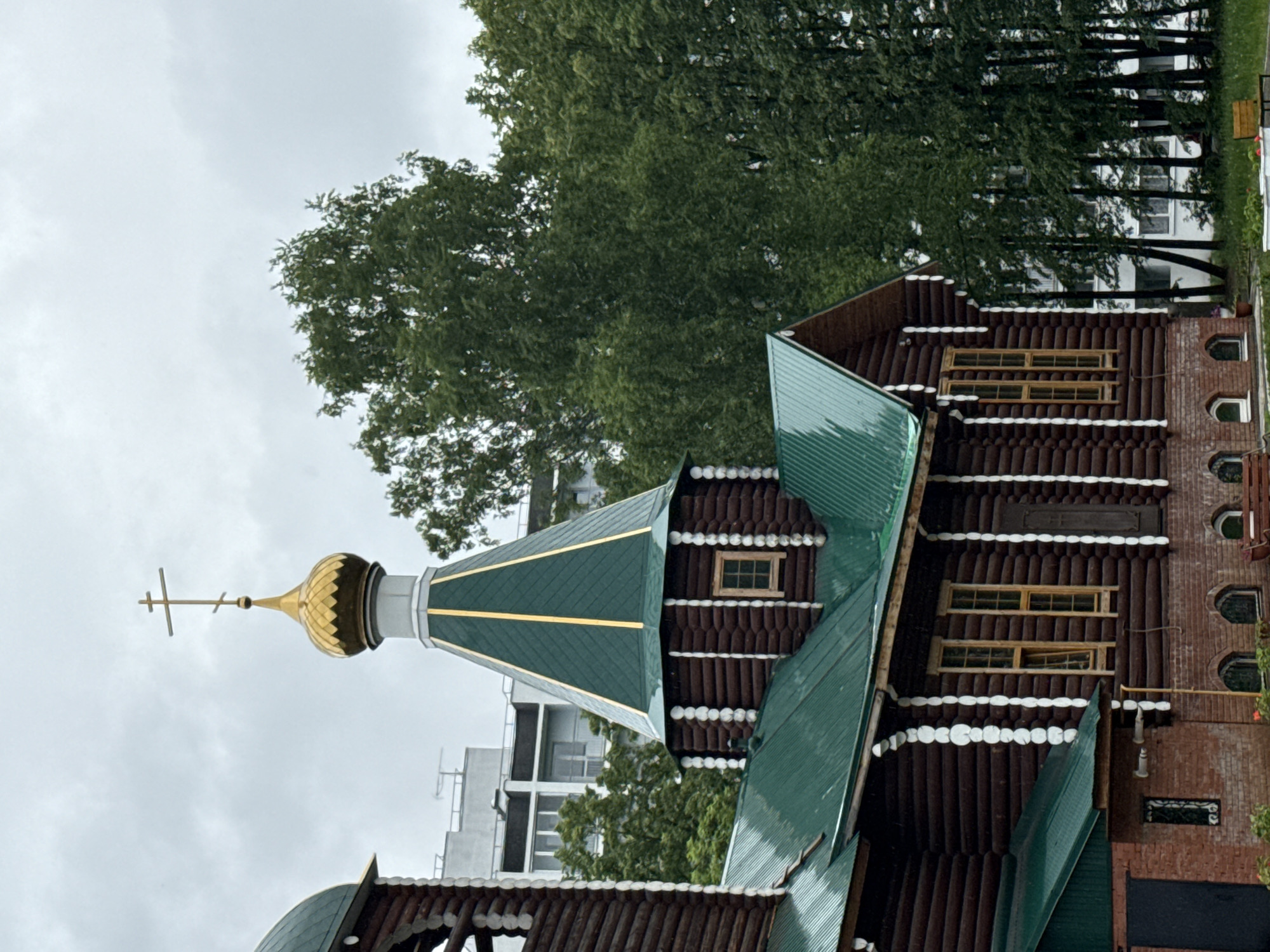 The image shows an Orthodox church in the city of Chaikovsky, Russia. The church has a golden dome with a cross on top, green roofs, and a brown wooden facade with white decorative elements. There are trees and buildings in the background. The sky is overcast.