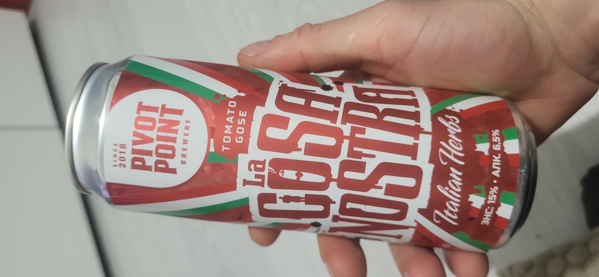 A person is holding a can of Pivot Point Brewery's "La Cosa Nostra" Tomato Gose beer. The can is red, white, and green, in the colors of the Italian flag. The words "La Cosa Nostra" are printed in large red letters on the can. The phrase "Italian Herbs" is also visible. The can also features the Pivot Point Brewery logo and indicates the beer is a Tomato Gose, and includes the alcohol content.