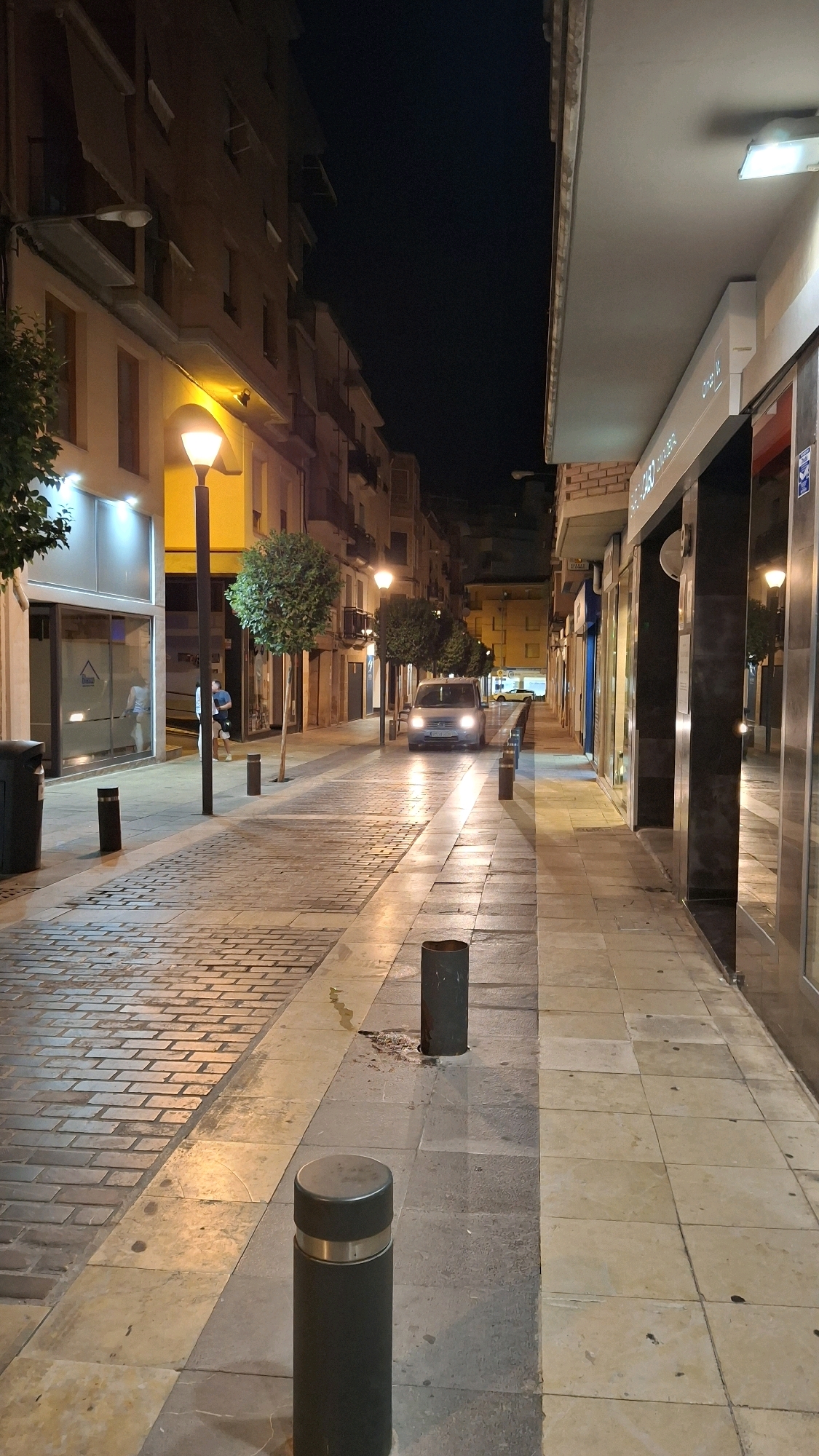 The image depicts a nighttime street scene in Alcañiz, Spain, specifically on Calle Blasco de Alagón. The street is illuminated by lampposts and the lights from storefronts. On the left side of the street, a building with multiple floors features balconies and awnings. A lamppost with a yellow glow is visible, as is a trash receptacle. A storefront with large windows is also present. Further down the street, trees are planted along the sidewalk. On the right side, a building houses commercial establishments with prominent glass facades. A silver van is driving down the street, its headlights illuminating the road. The street itself is paved with cobblestones and square tiles. Several black bollards are strategically placed along the sidewalk.