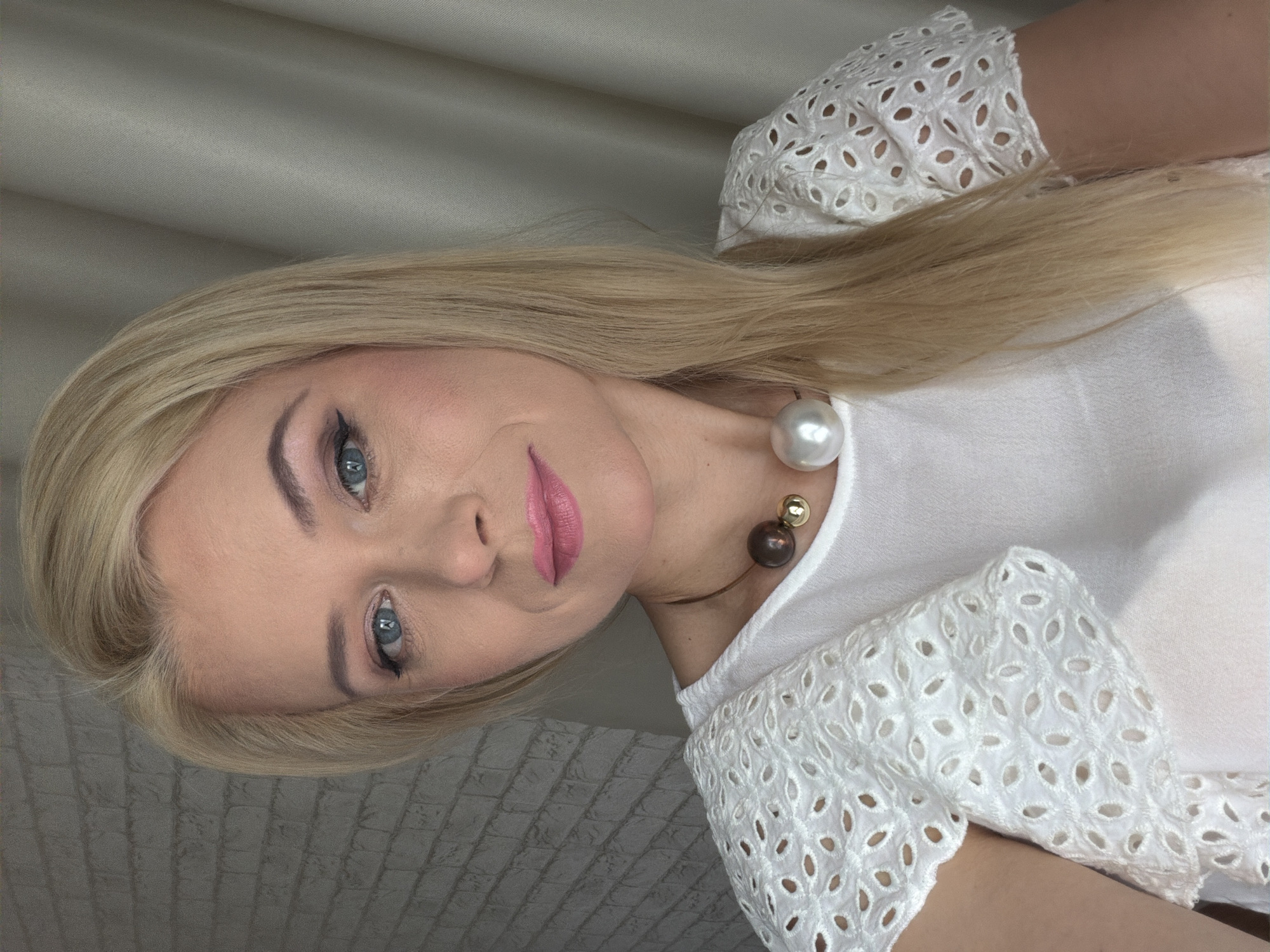 The media file is a portrait of a blonde woman with blue eyes and pink lipstick. She is wearing a white top with eyelet details on the sleeves and a pearl necklace. The background is a neutral-colored fabric. The woman is looking towards the camera with a slight smile. The image appears to be taken indoors. There is no visible text in the media file.