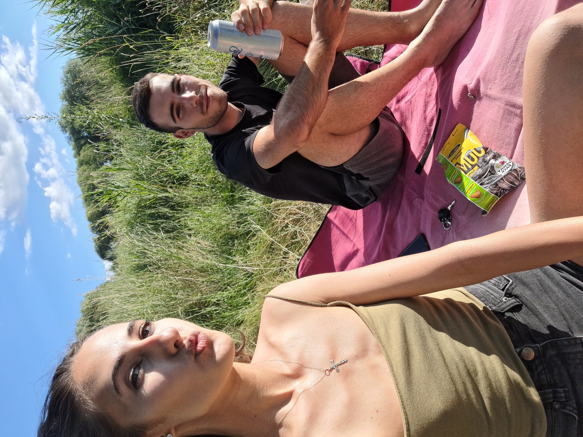 Two young adults are seated on a pink blanket in a grassy area, likely a park or field. A young woman in the foreground, facing left and making a pouting expression, wears a green tank top and black denim shorts. She has a cross necklace. A young man, partially visible in the background on the right, wears a black t-shirt and grey shorts. He is holding a silver can and looking towards the camera. A bag of snacks, car keys, and a cell phone are on the blanket between them. The background consists of tall green grass and trees under a bright blue sky with scattered clouds. The lighting suggests it is daytime, possibly late afternoon due to the long shadows.