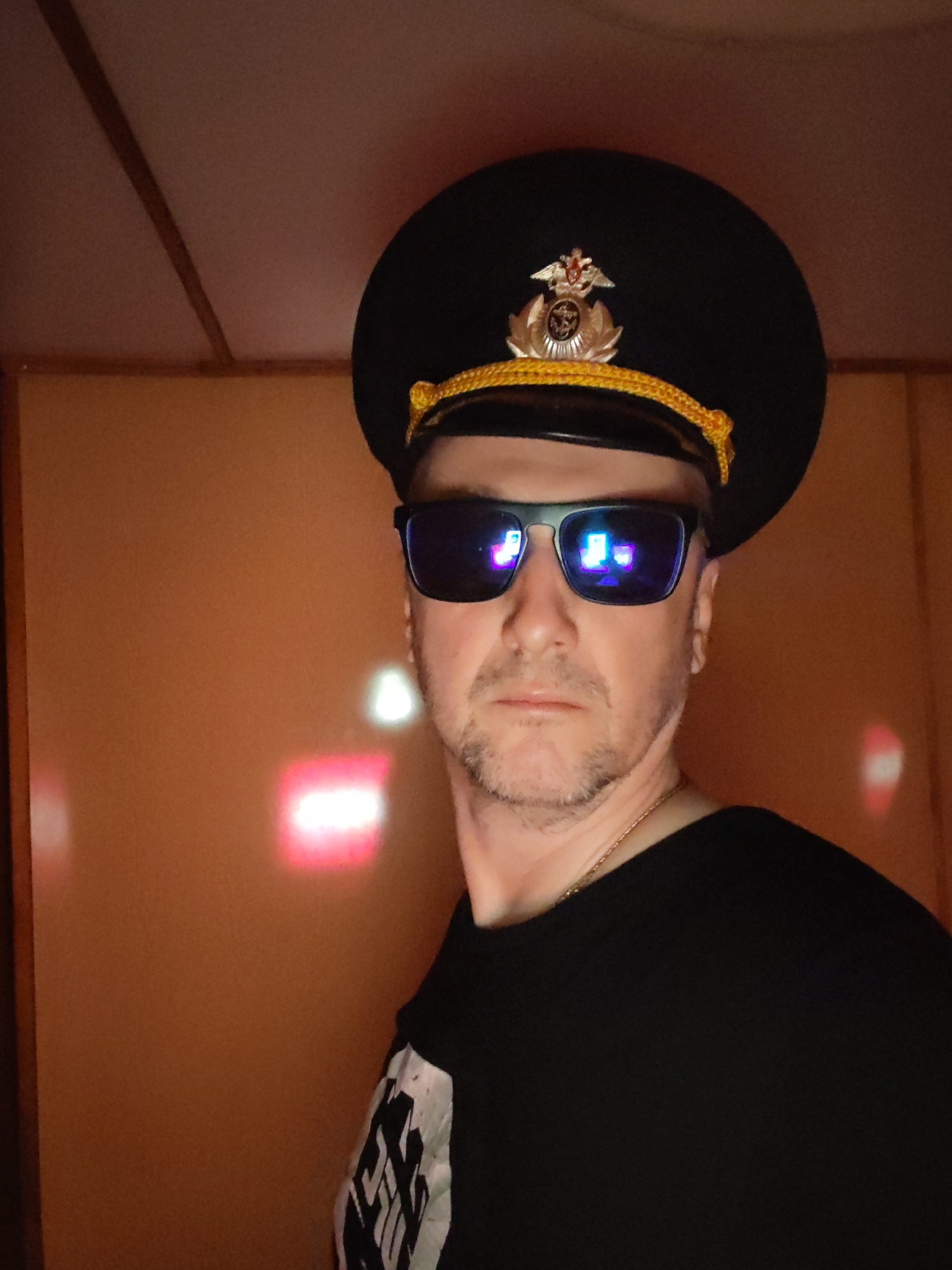 A man is depicted in a close-up, indoor portrait, seen from the chest up. He has a short beard and mustache and looks directly at the viewer. He wears a dark, military-style peaked cap adorned with a gold braided cord and a prominent gold and red emblem featuring a double-headed eagle with an anchor and wings, reminiscent of Russian insignia. His eyes are obscured by dark, reflective sunglasses that show distinct reflections of illuminated screens or digital displays. A thin gold chain is visible around his neck, and he is wearing a black short-sleeved t-shirt with an abstract white graphic design on the left sleeve and side.
The man is situated against a warm, light brown or orange-colored background that appears to be a paneled wall, extending to a similarly paneled ceiling with a visible seam. The lighting is dim, featuring prominent, blurry reddish-white glows on the wall behind him, which are also reflected in his sunglasses. The setting suggests an indoor environment, likely in the evening or at night given the reliance on artificial light. The man's expression is neutral and composed. No legible text is discernible on his clothing, hat, or in the reflections. The overall scene conveys an informal portrait. The location is Shcheleiki, Russia.