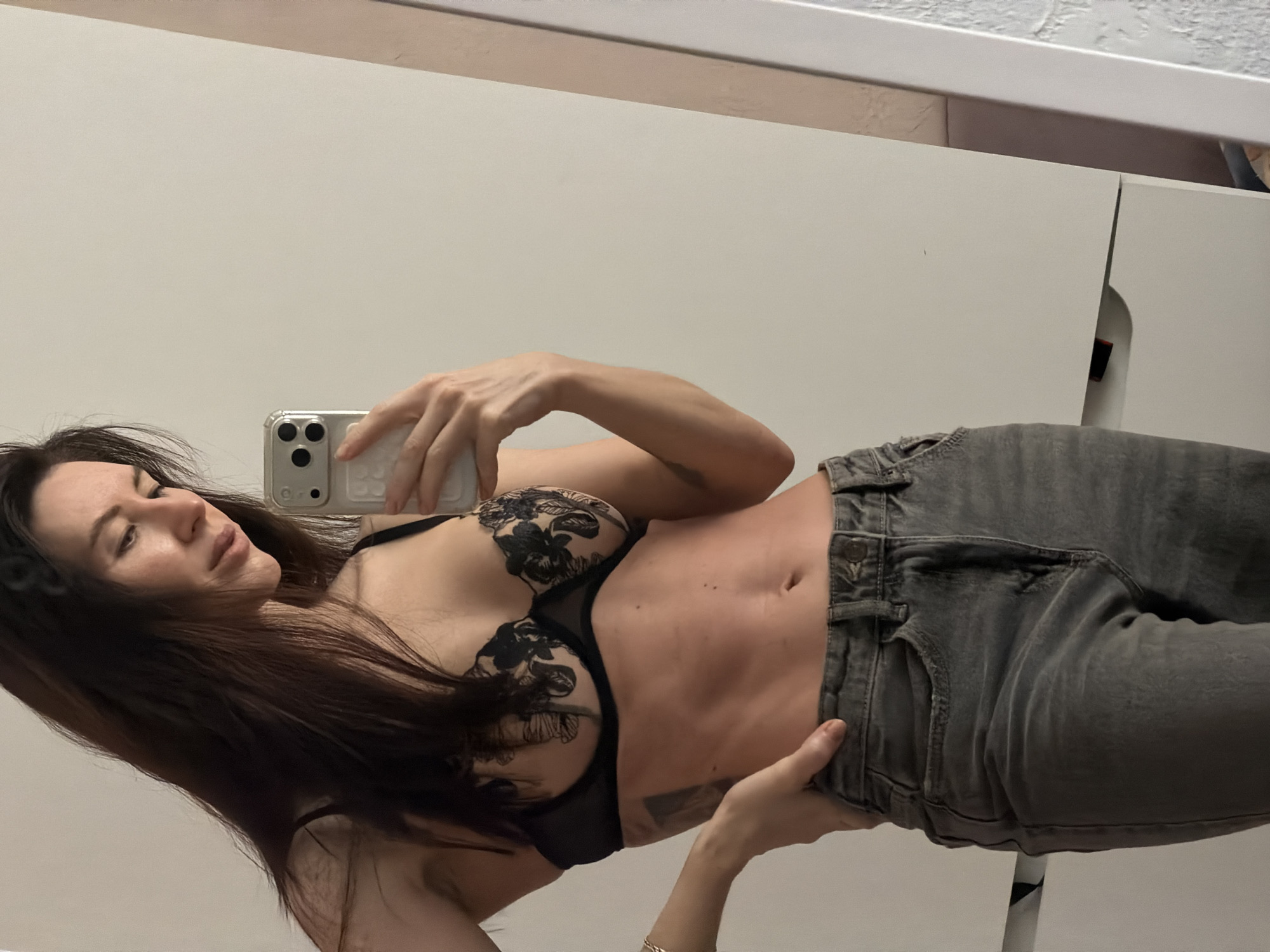 The image displays a tilted mirror selfie of a woman with long, dark hair. She is wearing a black lace bra with intricate floral patterns and dark grey jeans, which are partially unzipped and pulled down to reveal her toned midriff and navel. Her left hand holds a light-colored smartphone, likely an iPhone, directed towards her reflection to capture the image. Her head is tilted, and she looks towards the phone with a calm expression. The background consists of plain, light off-white wall panels or cabinet doors, with a thin horizontal trim visible along the top edge. A slight gap between the panels reveals a darker area behind. No discernible text is visible in the image.