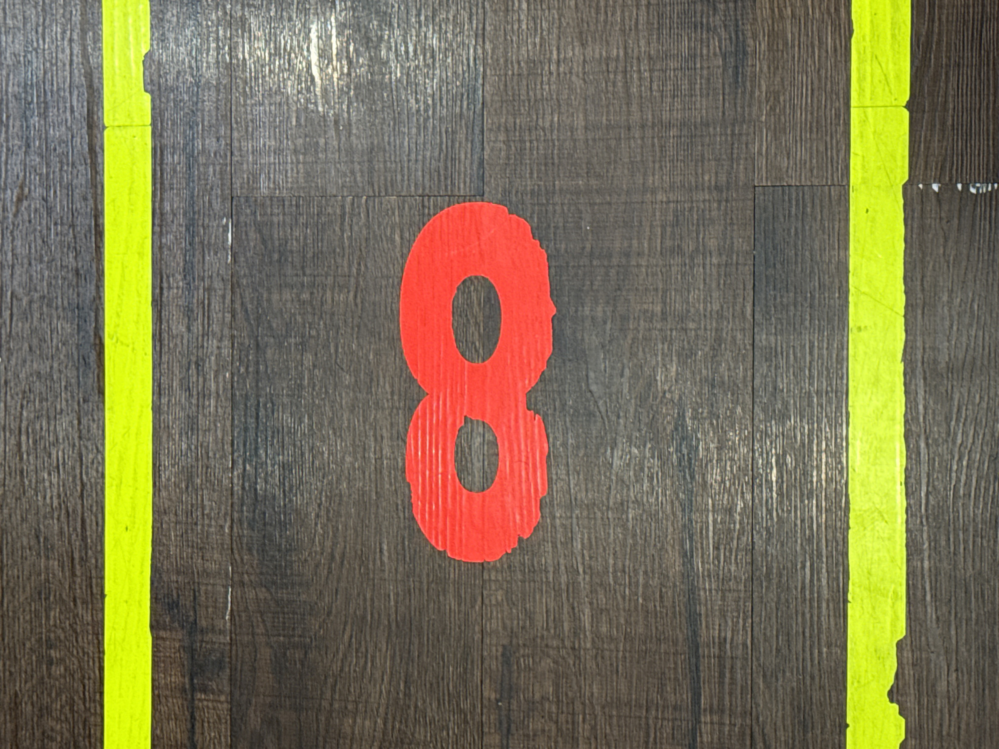 The image shows a close-up, top-down view of a wooden floor. Two bright yellow lines, likely tape, run horizontally across the image, one near the top and one near the bottom. In the center of the image, a red, paper-cutout shape resembling an infinity symbol (∞) is placed on the floor. The floor itself appears to be laminate or a similar material with a wood grain pattern. There are no people or other objects visible in the frame. The scene is devoid of any specific Da Lat city landmarks or context.