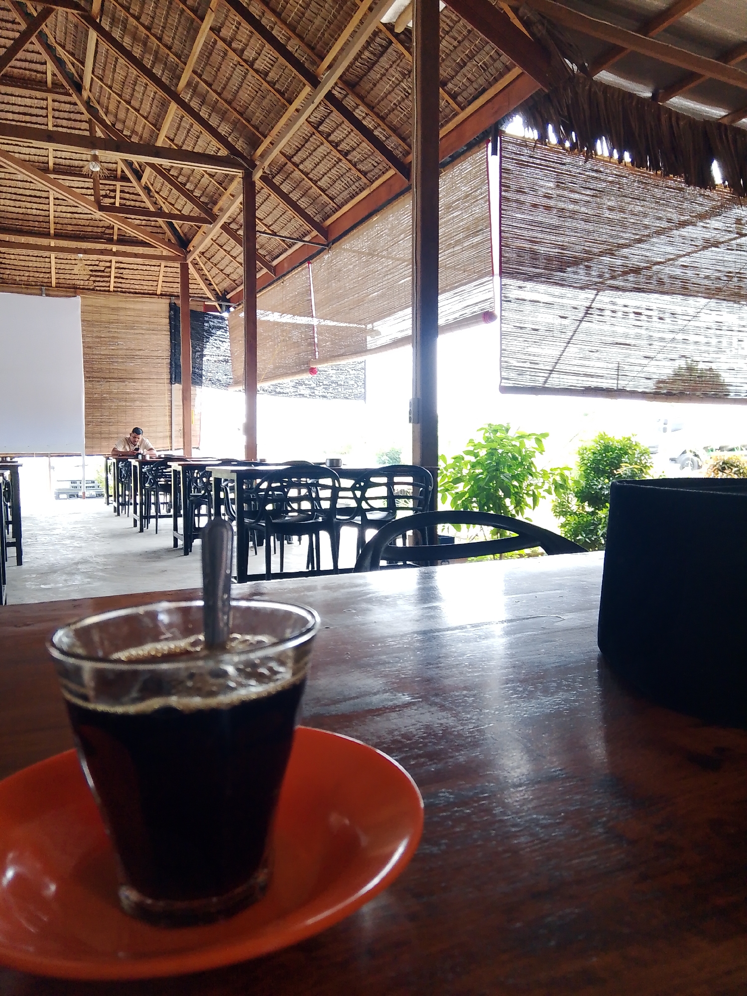In the city of Meunasahmee, Indonesia, this image depicts an indoor cafe with an open-air feel. The architecture features a thatched roof supported by wooden beams, with bamboo blinds covering the open sides. In the foreground, a glass of dark coffee sits on an orange saucer on a wooden table. A spoon is visible in the coffee. In the background, rows of tables and chairs are set up, and a lone person is seated at one of the tables, looking at their phone. Green foliage is visible outside the cafe, suggesting a lush environment. The lighting is natural, coming from the open sides of the cafe.