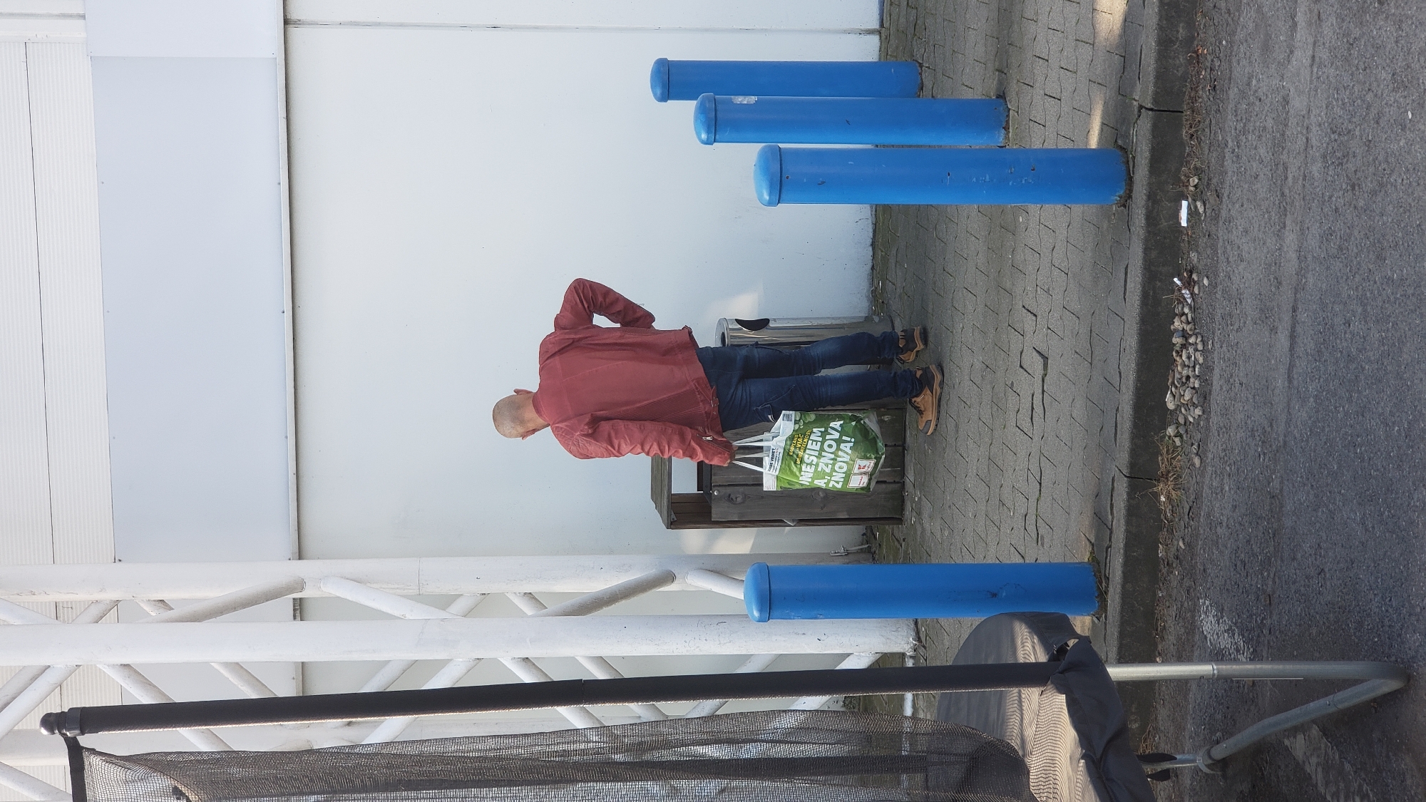 A bald man wearing a red jacket and blue jeans is standing next to a trash receptacle and a shopping bag. The man is holding the bag which has green lettering and a picture of fruit. The shopping bag is a reusable grocery bag. The man has his right hand on his hip. The trash receptacle is made of wood and metal. The scene is outdoors, on a paved area with blue bollards in the foreground. A white wall is in the background. This scene is located in Prešov, Slovakia, near a shopping mall.