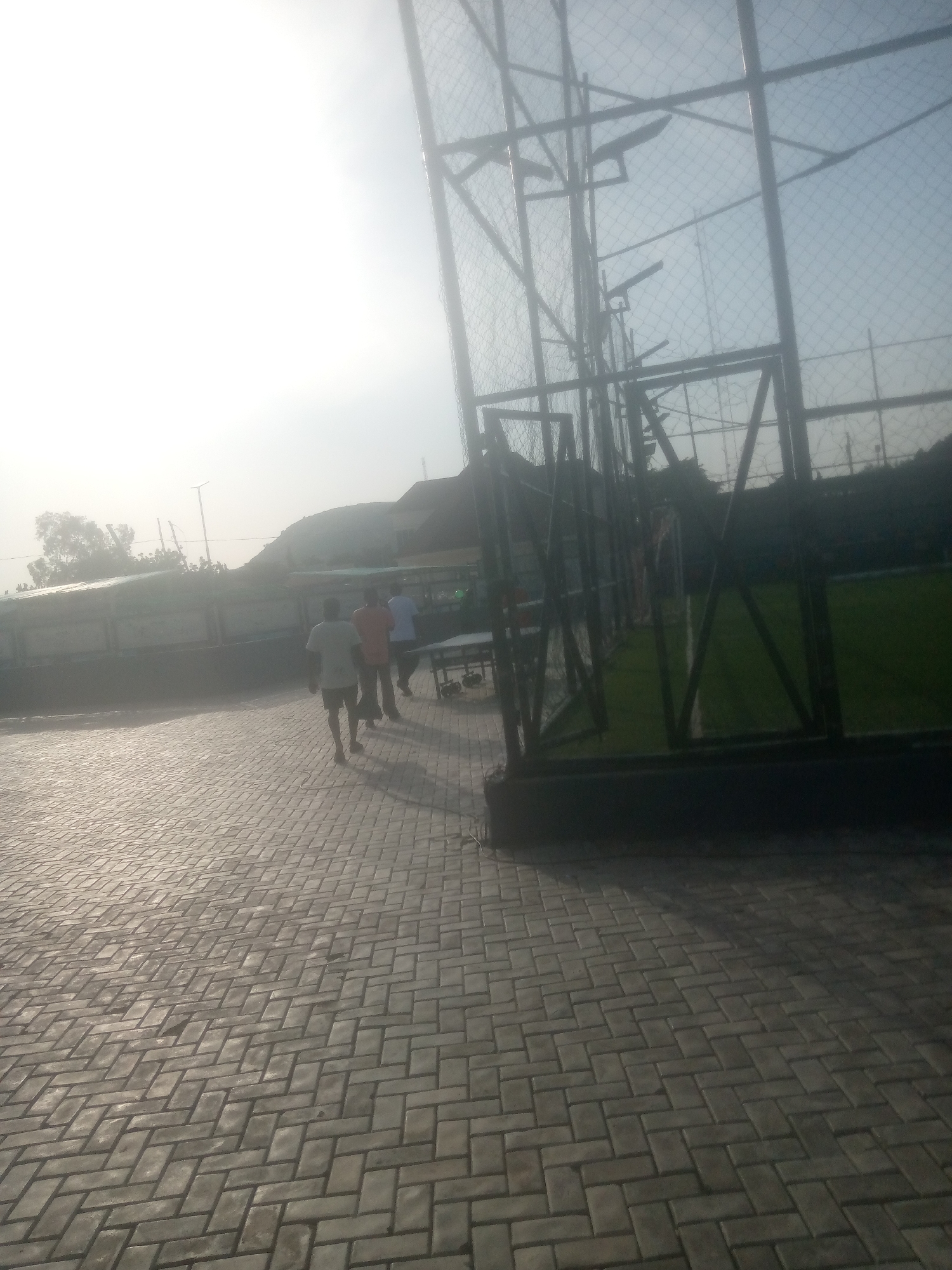 The image depicts an outdoor scene with a paved walkway in the foreground, laid with rectangular bricks in a herringbone pattern. The walkway is illuminated by natural light, suggesting it is daytime. In the middle ground, three individuals are walking away from the camera. The person in the front is wearing a white t-shirt and dark shorts, the second person is in an orange t-shirt, and the third person is in a blue t-shirt. Behind the people, there is a fenced sports field with artificial turf, indicating a possible sports facility. A large metal structure with netting, likely part of the sports field enclosure, dominates the right side of the image. In the background, there are buildings and vegetation, with a slightly hazy sky suggesting it might be early morning or late afternoon, or possibly overcast conditions. The overall impression is a casual, everyday scene at a sports complex. There is no visible text in the image.