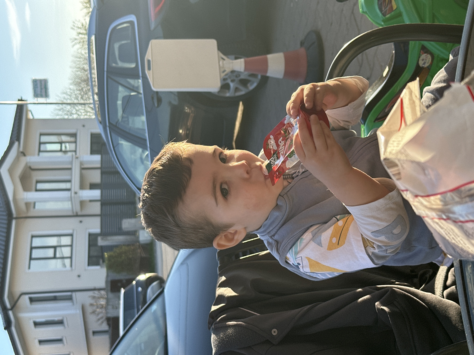 A young child is drinking from a red fruit puree pouch while seated in a stroller, facing away from the camera. The child has short dark hair and is wearing a light-colored long-sleeved shirt under a grey vest. A black jacket or blanket is draped over the stroller seat. Behind the stroller, part of a black car is visible, with a white and orange traffic cone nearby. A modern building with multiple windows is situated across the street, along with a street sign. The location is likely in Plovdiv, Bulgaria, although no details uniquely confirm this.