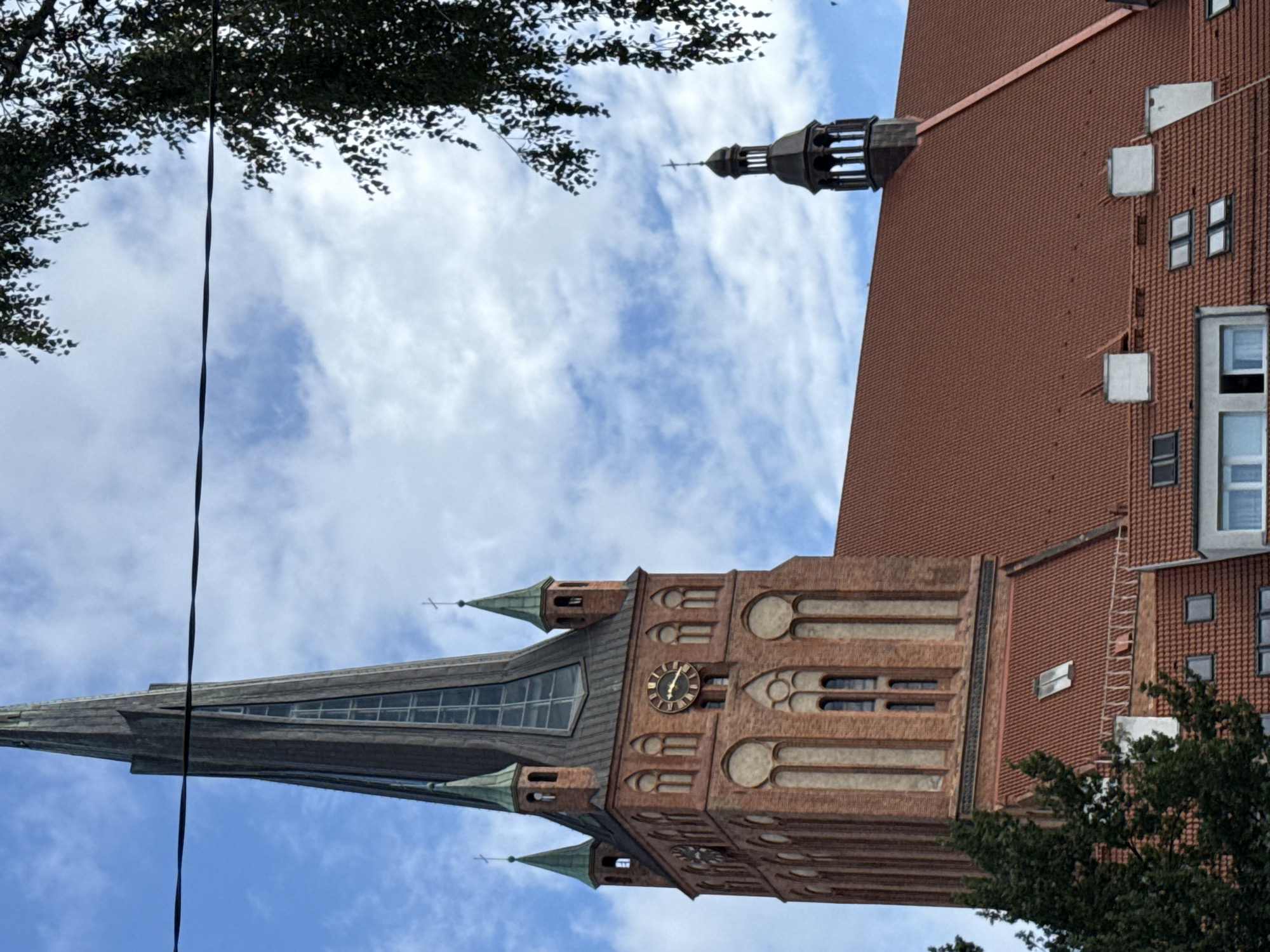 The image captures a view of the St. James' Church (Kościół św. Jakuba Apostoła) in Szczecin, Poland. The prominent brick tower of the church with a tall, dark spire dominates the center of the frame, reaching towards a partly cloudy blue sky. A clock face is visible on one side of the tower. Smaller, green-capped turrets flank the main spire. A red-tiled roof extends from the base of the tower to the right, with a smaller dark tower topped by a cross visible further in the distance. The lower portion of the image shows a glimpse of surrounding buildings with red tile roofs and some trees on the left. A dark wire runs horizontally across the upper part of the image.