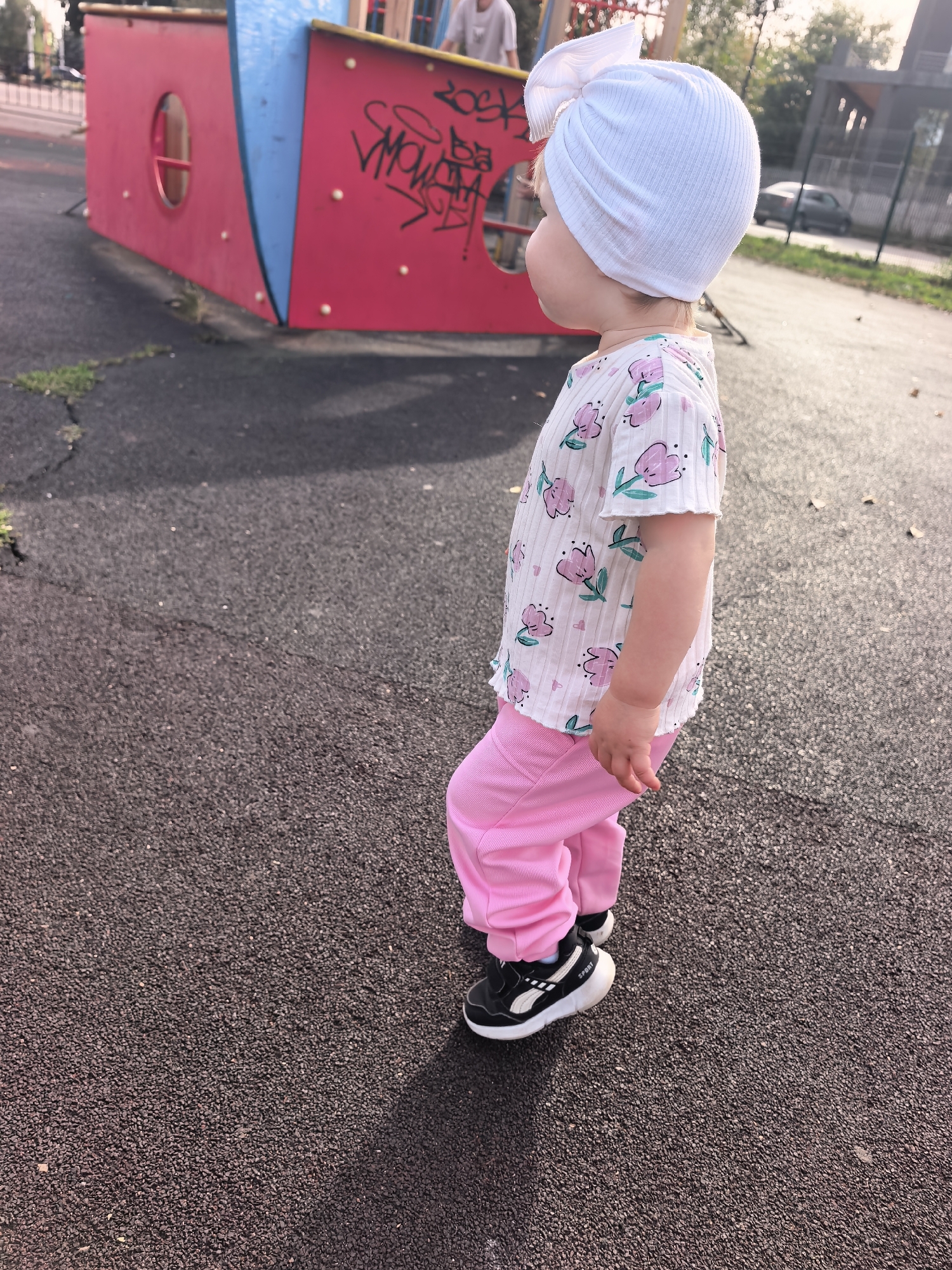 A child, viewed from the side, stands on a paved playground surface, facing right. The child is wearing a white ribbed t-shirt with a pink floral print, pink pants, a white turban with a large bow, and black and white sneakers. Behind the child, a red and blue playground structure is visible, with graffiti on its red surface. In the background, a fence and trees can be seen, with a car driving by on a road. The lighting suggests it is daytime, with shadows cast on the ground. The location is identified as a playground in Sterlitamak, Russia.