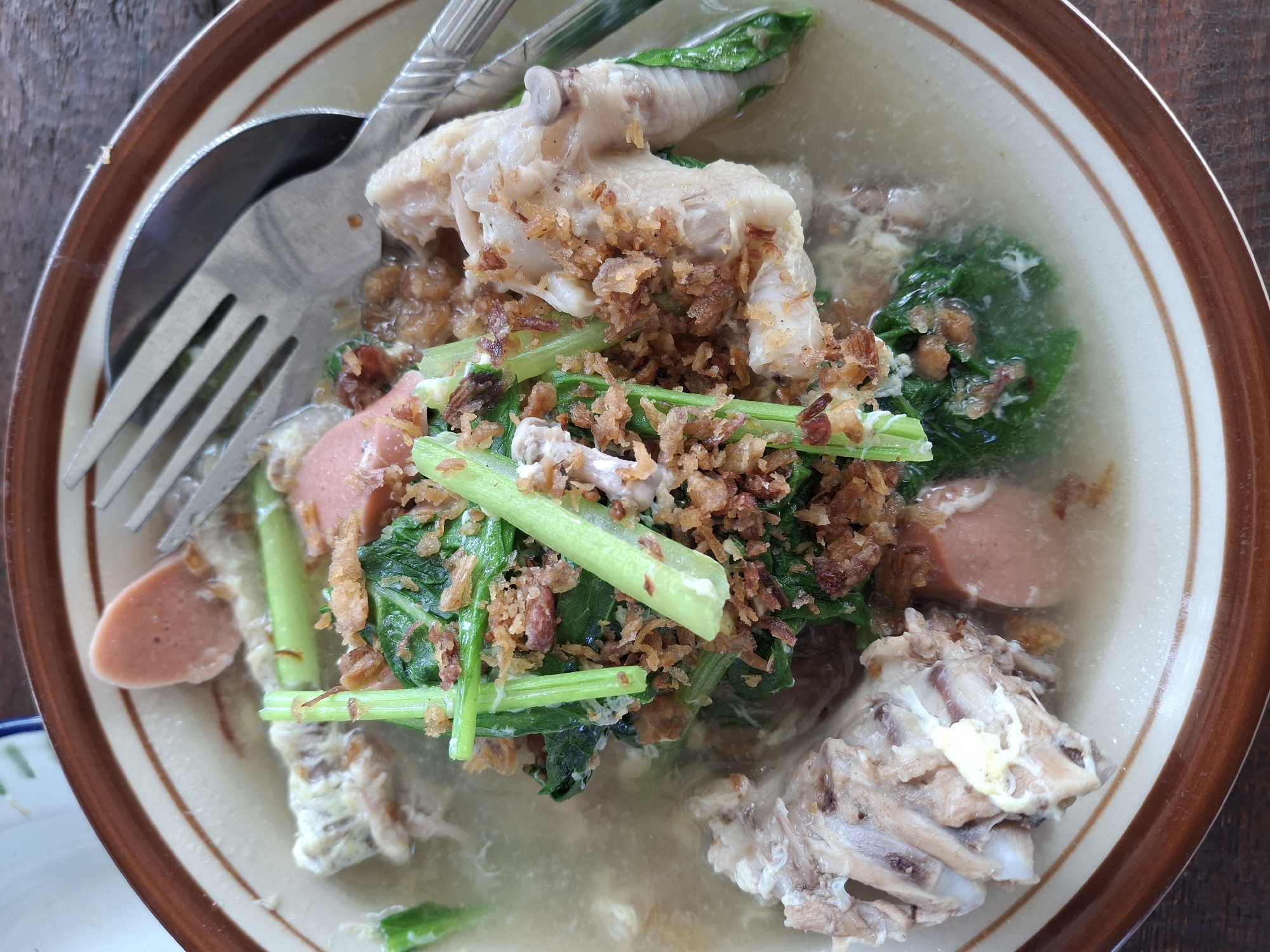 A close-up overhead shot shows a bowl of soup containing bone-in chicken pieces, sliced sausages, leafy green vegetables, and fried shallots. A fork and spoon are resting on the edge of the bowl. The bowl is set on a dark wooden surface.