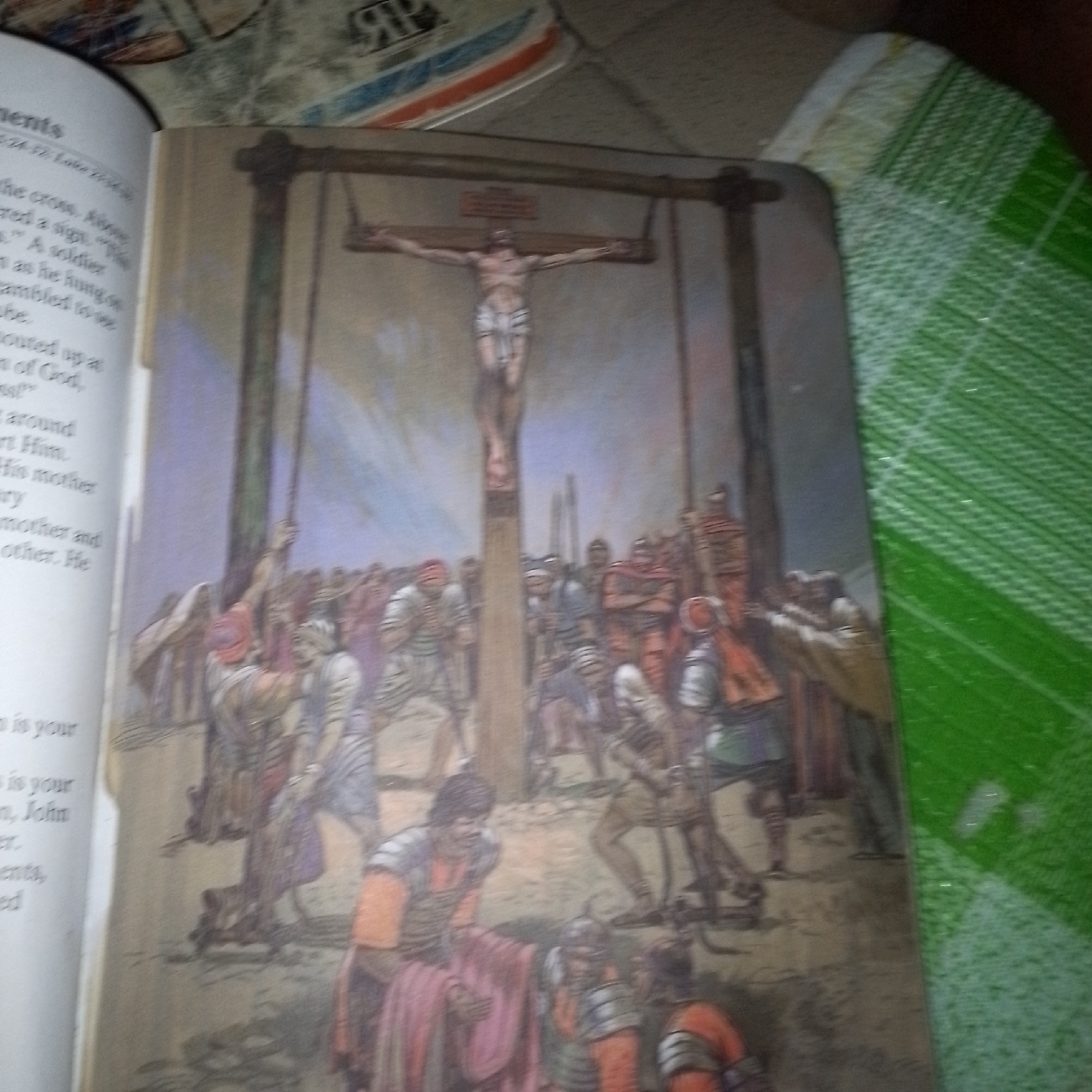 The image shows a painting of the crucifixion of Jesus. Jesus is depicted on the cross, with a crowd of people gathered below. Some of the people appear to be soldiers, while others are onlookers. The scene is set in what appears to be an outdoor location, with a rocky landscape and a cloudy sky. The painting is detailed, with a focus on the emotional expressions of the figures. The image also shows a book with text in English on the left side, and a green checkered fabric on the right side.