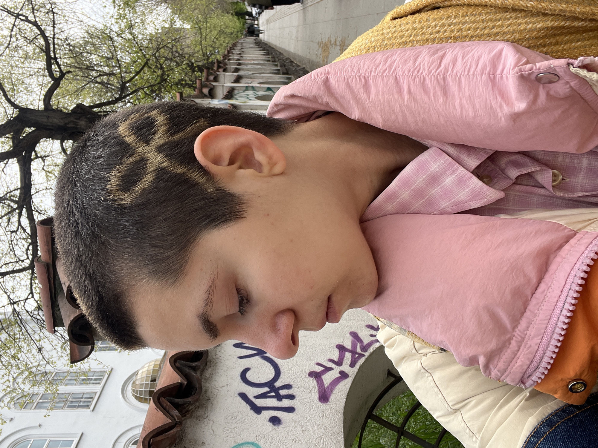 A close-up shot from the side shows a person with short, dark hair, featuring a patterned design shaved into it. The person is wearing a pink shirt and jacket. The background features a white building with arched windows and the word "JACK" graffitied on it, along with greenery. The person appears to be near the Spanish Steps in Belgrade, Serbia.