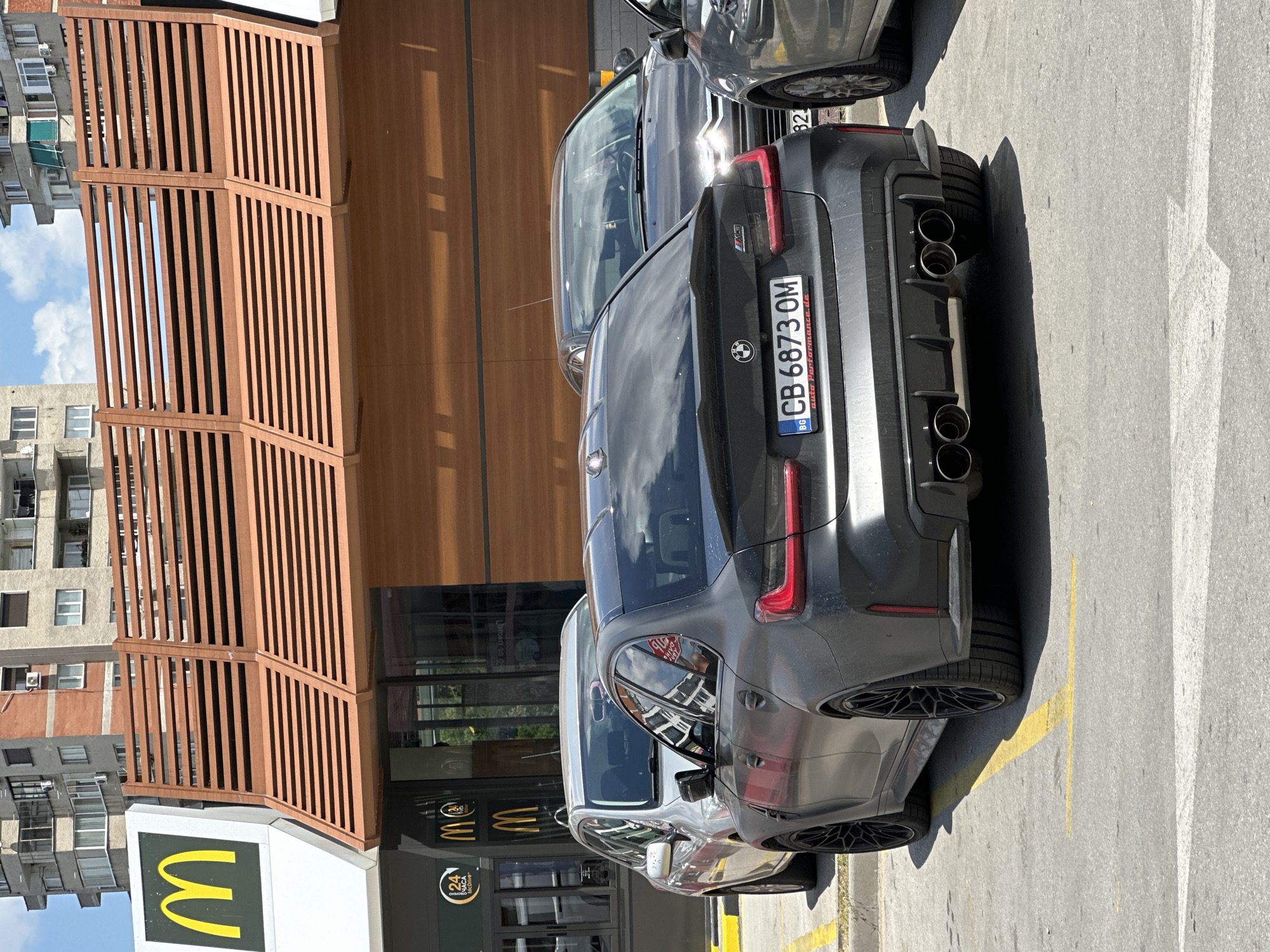 A dark grey BMW M3 sedan is parked in a McDonald's parking lot in Veliko Tarnovo, Bulgaria, on a cloudy day. The car, facing away from the camera, has a license plate that reads "CB 6873 OM". The McDonald's building behind it features a large golden arch logo and a wooden slatted facade. Another car, a silver sedan, is parked to the left of the BMW. The parking lot is paved with asphalt and marked with yellow parking lines. In the background, apartment buildings are visible.