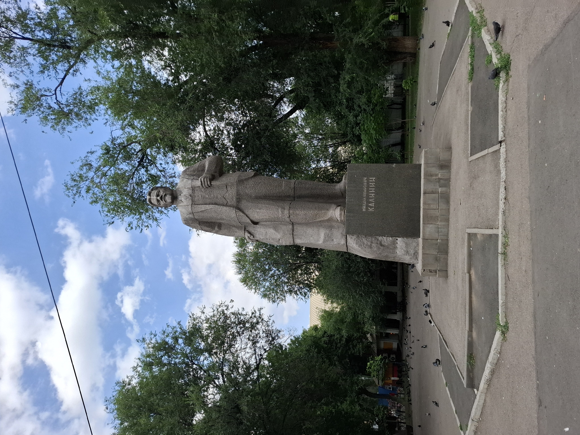 The image shows a full-body statue of a man, likely Mikhail Kalinin, a Soviet statesman. The statue is made of stone and depicts him in a suit, standing with one hand resting on his chest and the other on his hip. The statue is mounted on a rectangular pedestal, which has text inscribed on it in Cyrillic. The text appears to read "МИХАИЛА ИВАНОВИЧА КАЛИНИН".
The statue is situated outdoors, with lush green trees and a bright blue sky with scattered white clouds in the background. The ground around the statue is paved with asphalt, with concrete borders and some patches of grass. Several pigeons are scattered on the ground and the paved areas, suggesting a public space. The overall impression is that of a monument in a park or public square. The lighting and the presence of green foliage suggest it is daytime, likely late spring or summer. The image is captured from a low angle, looking upwards towards the statue and the sky. There are no people visible in the immediate vicinity of the statue, but further in the background, on the left side, there are some buildings and what appears to be a street with some parked bicycles and possibly market stalls. The overall mood is peaceful and serene.