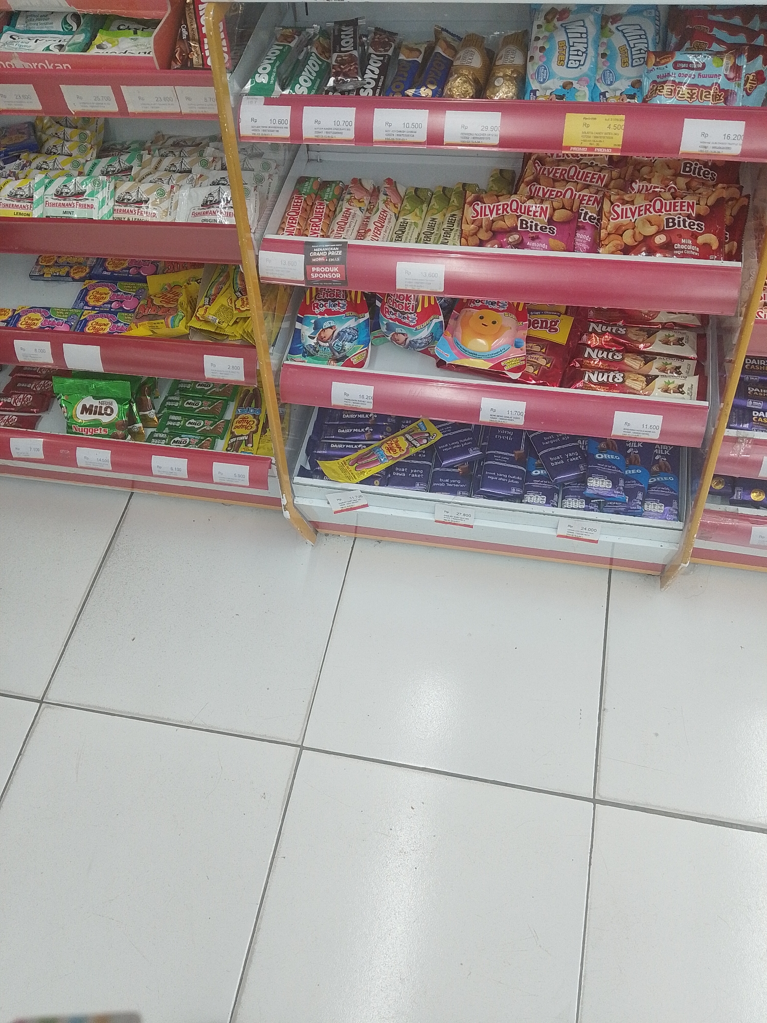 The image shows a close-up, eye-level, medium shot of shelves stocked with various snacks in a retail environment. The lighting appears to be from overhead fluorescent fixtures, casting a neutral white light on the products. The shelves are red, with white price tags visible on the lower shelves. The background consists of white tiled flooring.
The products displayed are predominantly confectionery and snacks. On the top shelf, several brands of chocolates are visible, including what appear to be Ferrero Rocher chocolates, a pack of "Milka" branded chocolates, and other assorted chocolate bars.
The second shelf from the top features rows of "Fisherman's Friend" lozenges in various flavors like Lemon and Mint, as well as a shelf of "Silver Queen" chocolate bars, specifically "Bites" in varieties such as Almonds and Milk Chocolate Cashew.
The third shelf displays "Chupa Chups" lollipops, a "Choki Choki" snack, and several packages of "Silver Queen" chocolates. Towards the right, there are multiple bars of "Oreo" biscuits.
The bottom shelf exhibits "Nestle Milo" nuggets, "Kit Kat" bars, and what appears to be "Dairy Milk" chocolate bars. The price tags indicate that the items are priced in Indonesian Rupiah (Rp).
There are no people visible in the image. The focus is solely on the organized display of packaged food items. The overall impression is of a well-stocked convenience store or supermarket aisle. The image is taken in Uwuran Satu, Indonesia.