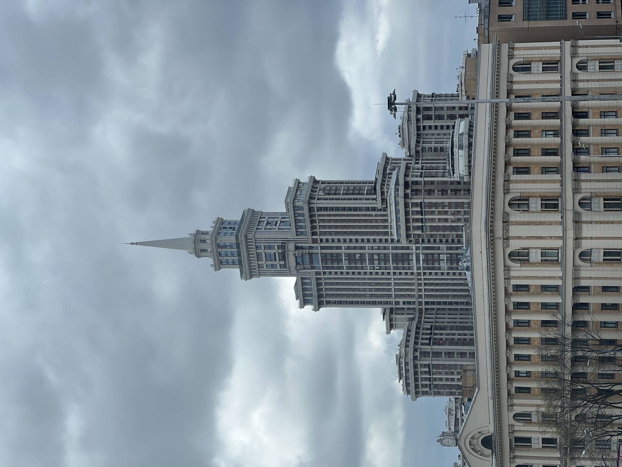 The image features a tall, multi-tiered building under a cloudy sky. The building has a spire at the top. Adjacent to this building is another structure with a different architectural style, featuring rows of windows and columns. The location is identified as Khimki, Russia.