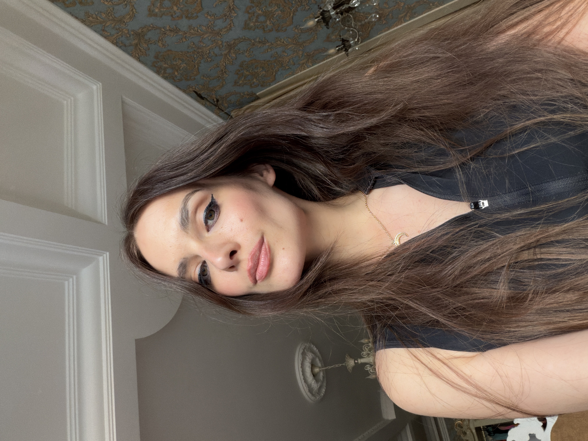 A close-up, rotated portrait shows a young woman with long, dark brown hair that cascades around her shoulders. She is wearing a black top with a zippered neckline and a delicate gold necklace with a crescent moon pendant. Her makeup includes winged eyeliner and a soft pink lipstick. The background features ornate wallpaper with a damask pattern and a vintage chandelier. To the left, a white door with raised paneling is visible. The lighting suggests it is daytime, and the overall ambiance is intimate and perhaps taken indoors. The image appears to be a selfie, with the woman looking towards the camera.