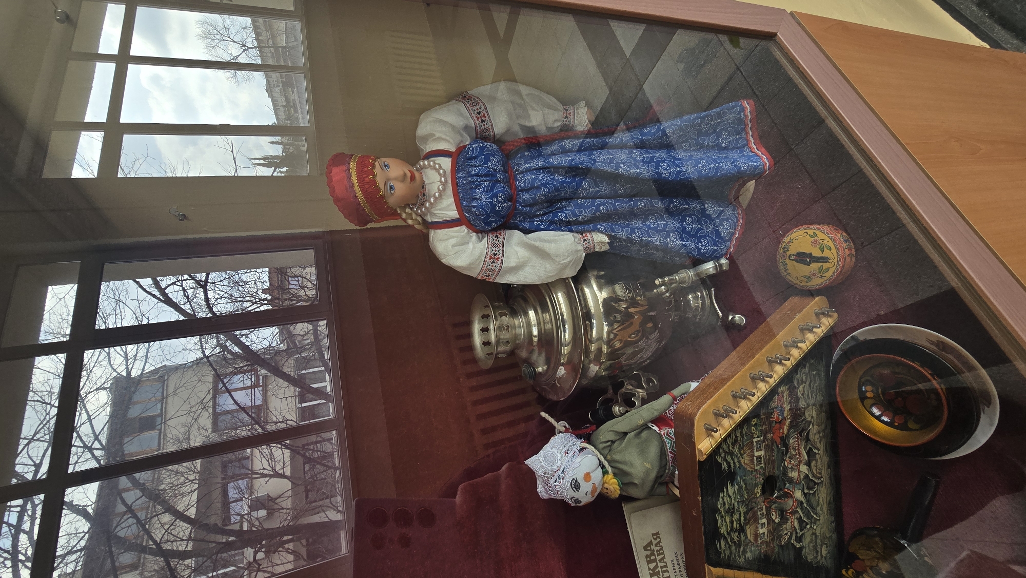 A display case at the Regional History Museum in Stara Zagora, Bulgaria, contains a Russian doll dressed in traditional blue and white clothing, a samovar, a painted wooden box, a gusli (a Russian stringed instrument), and other traditional Russian artifacts. The display case is enclosed in glass, reflecting the outside environment, including a nearby building and bare trees. The interior of the case is lined with a red velvet material.