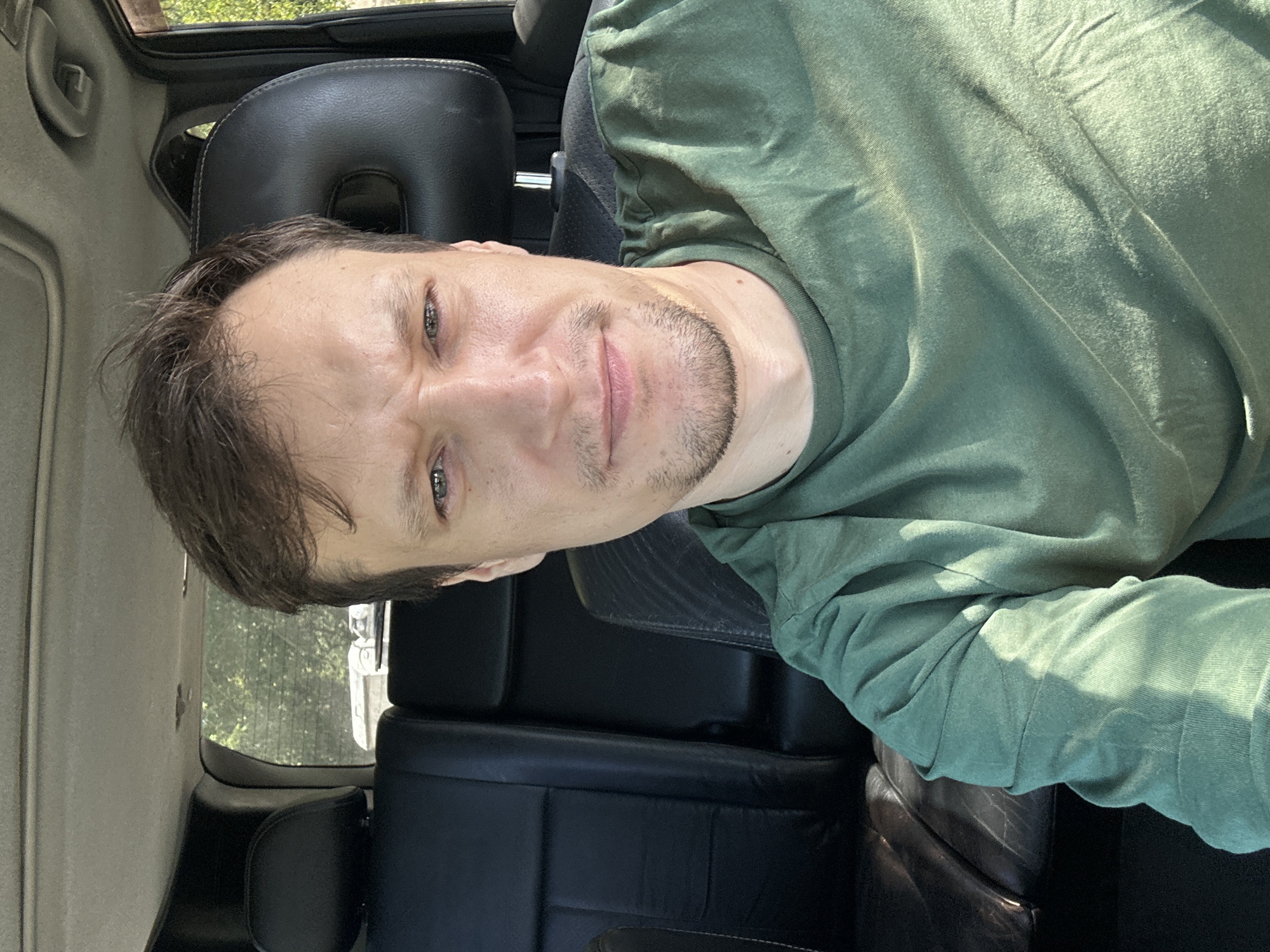 A man is seated in the driver's seat of a vehicle. He is wearing a green t-shirt and has short brown hair and a light beard. The interior of the car is visible, including black leather seats and the dashboard. Sunlight streams in from the right side of the image, casting shadows on the man and the interior. The background outside the car appears to be trees and foliage, suggesting a rural or park-like setting within or on the outskirts of Makiv, Ukraine.