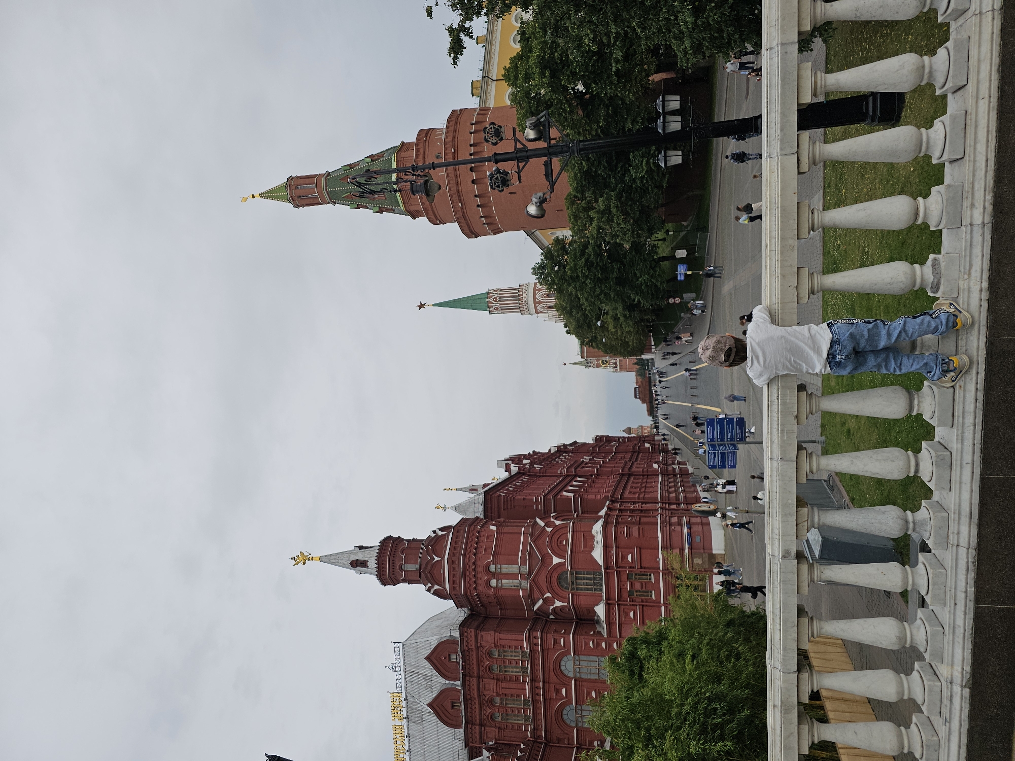The image shows a child, viewed from behind, leaning on a white marble balustrade and looking out at a street scene in Moscow, Russia. The child is wearing a light-colored t-shirt, blue jeans with a design on the leg, and a baseball cap.
In the background, the street is lined with people walking. On the left side of the street is a large, red brick building with white trim and ornate architecture, identified by text on the roof as the "State Historical Museum." On the right side of the street, several towers are visible. One prominent tower is round and made of red brick, topped with a green, pyramidal roof and a golden spire. Further down the street, another tall, slender tower with a green roof and a red star on top is visible. Trees and lampposts are scattered throughout the scene. The sky is overcast. The perspective suggests the viewpoint is from an elevated position, possibly a bridge or a higher level of a building, overlooking the street.