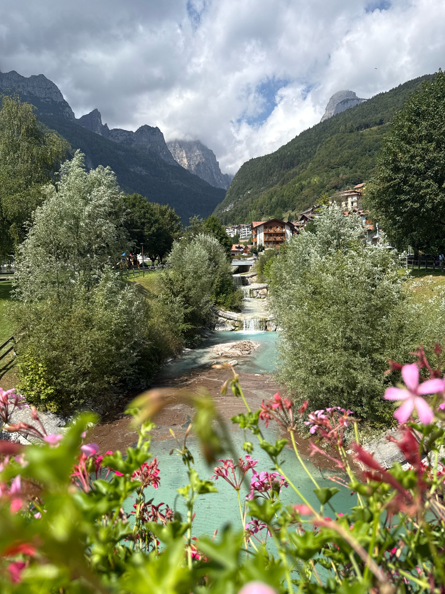 The image depicts a scenic view of a village nestled in a valley with mountains in the background. A vibrant turquoise river flows through the foreground, bordered by lush greenery and trees. In the mid-ground, a small village with traditional European architecture is visible, with houses featuring terracotta roofs. The river cascades over a series of small waterfalls as it winds through the village. The mountains are steep and rugged, with some peaks shrouded in clouds. The sky is partly cloudy, with patches of blue sky peeking through. In the immediate foreground, out-of-focus pink and red flowers add a pop of color to the scene. The overall impression is one of natural beauty and tranquility. The location is the town of Molveno, Italy, known for its picturesque setting in the Dolomites.