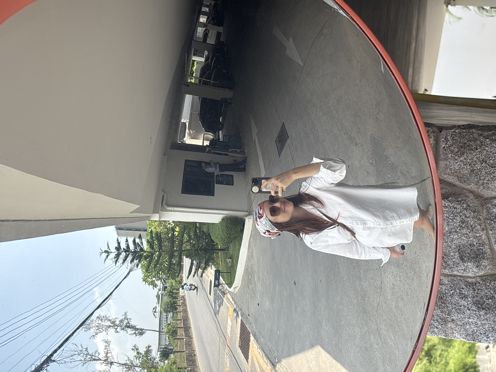 A full-shot photograph depicts a woman taking a mirror selfie in a large, circular convex security mirror set in an outdoor environment in Choeng Thale, Thailand. The image is rotated 90 degrees counter-clockwise.
The central subject, a woman, is reflected prominently in the mirror. She has long, dark brown hair, wears sunglasses, and a patterned headscarf with red and white tones. Her attire consists of a loose-fitting white long-sleeved dress or tunic. She holds a smartphone with multiple camera lenses up to take the selfie, and in her left hand, she holds a clear plastic water bottle. Her expression is neutral.
The convex mirror distorts the reflection, providing a wide-angle view of the surroundings. Inside the mirror's reflection, behind the woman, is a grey concrete or asphalt surface with yellow and white road markings, including white directional arrows and a section of yellow curb. A drain grate is also visible on the ground. Further back in the reflection, several parked cars are visible near a multi-story building entrance or parking area. A person wearing a light shirt and dark pants stands near this entrance. Green foliage, including trees and bushes, and a clear blue sky with some clouds are also reflected.
Outside the mirror's frame, on the left side of the image, the actual view shows more blue sky, utility poles with numerous wires, and distant buildings. On the right side, the mirror's reddish-orange frame is visible, and the texture of a stone wall or barrier. Further down the road, outside the mirror's view but visible in the original image, a person on a scooter or motorcycle is driving. A white sign with the number "1" and possibly an "S" next to it is visible by the roadside.
The scene suggests a sunny daytime setting due to the bright light and distinct shadows. The overall impression is of an informal outdoor moment captured through the unique perspective of a convex mirror.
