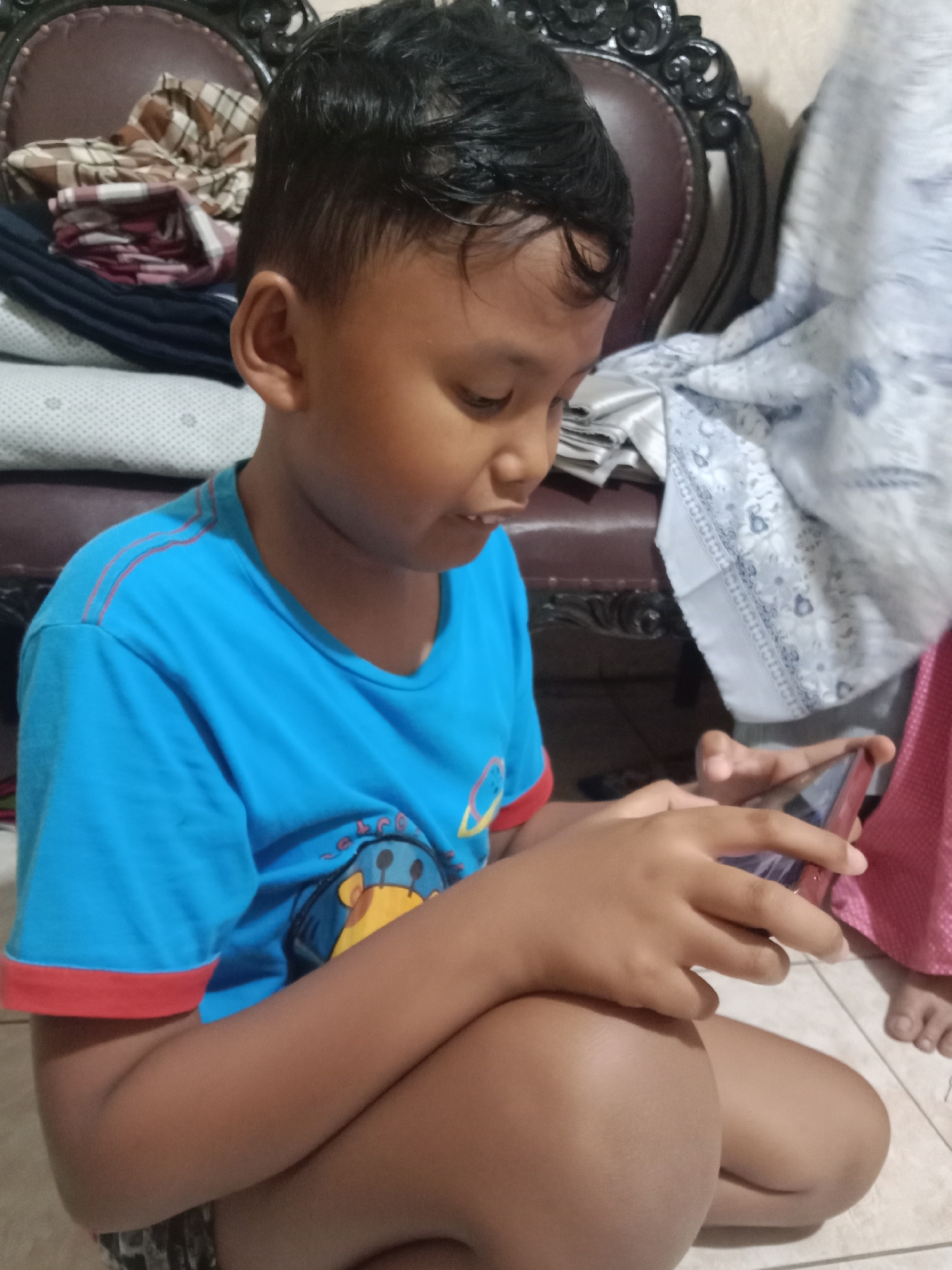 A young boy, likely of Asian descent, is shown sitting cross-legged and playing a mobile game on a smartphone. He is wearing a bright blue t-shirt with red trim on the sleeves and a design on the chest featuring a cartoon giraffe. The background includes stacked fabrics, possibly bedding or clothing, and a dark, ornate chair. Another person's legs are visible to the right, wearing a pink garment. The flooring appears to be tiled. The scene is indoors, and while the specific location in Madiun, Indonesia, is not identifiable from the image alone, it depicts a common domestic setting.