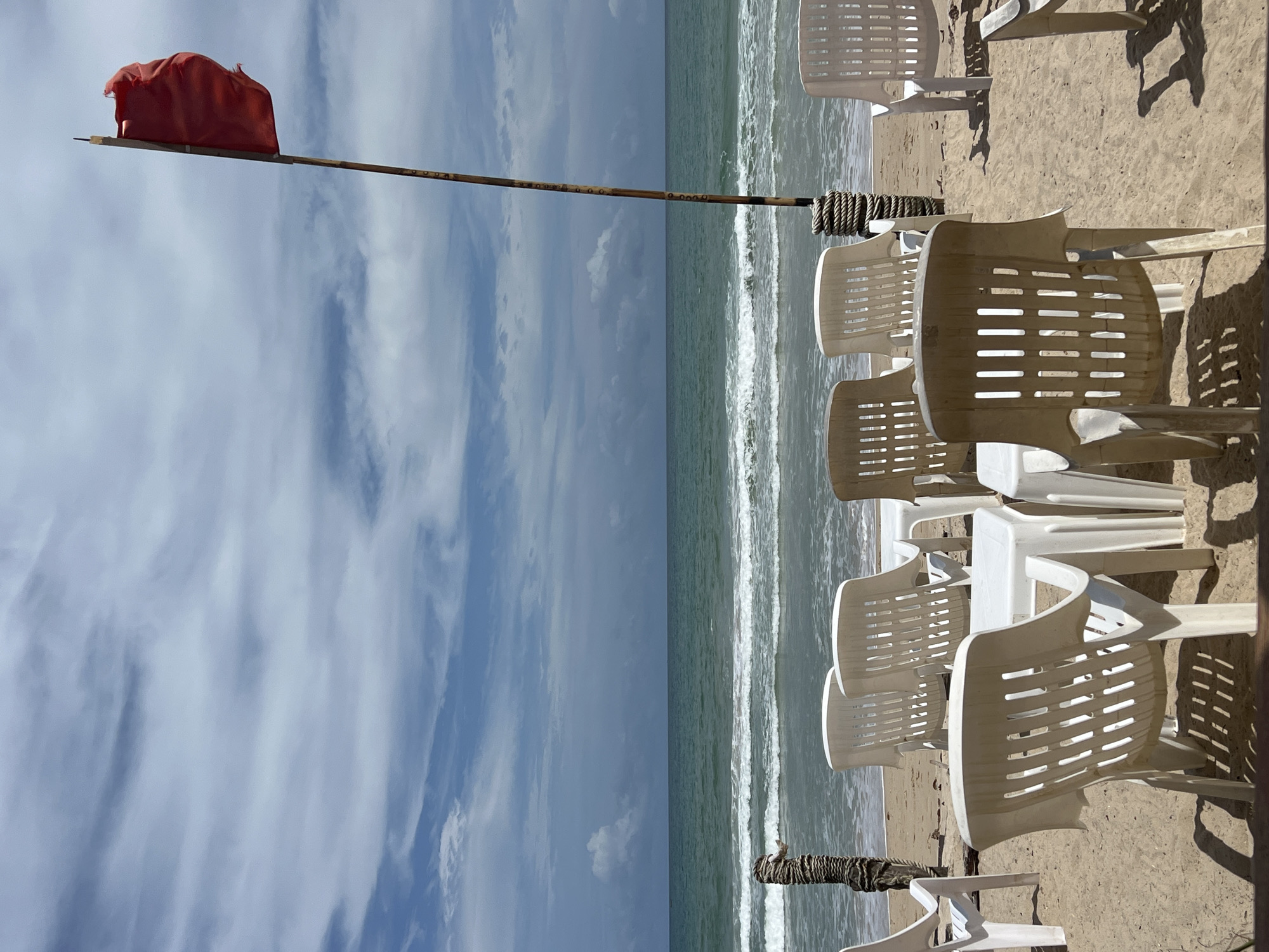 An outdoor scene depicts a beach with several white plastic chairs and small white tables arranged on the sand. In the background, the ocean stretches to the horizon under a sky with scattered clouds. A tall wooden pole stands to the right of the frame, topped with a rippling red flag. The overall impression is of a deserted beach setup, possibly for a cafe or rental service.
