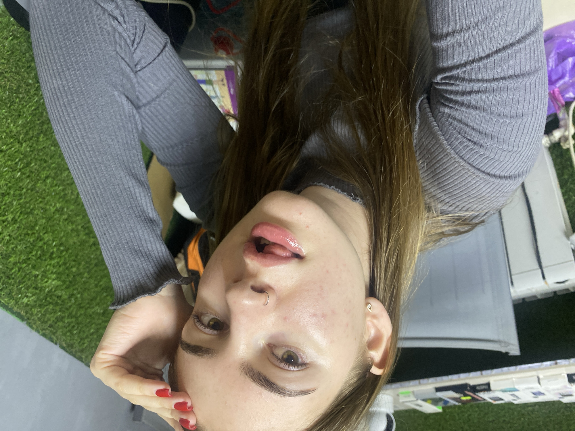 A young Caucasian woman with long, brown hair and light skin is taking a selfie. She is wearing a grey, ribbed turtleneck sweater. Her left hand is resting on her forehead, and her fingernails are painted red. She has a nose ring and a small stud earring in her left ear. Her mouth is slightly open, and her tongue is visible. The background appears to be a room with artificial grass on the right and shelves with various items on the left. There is also a white radiator or air conditioner unit visible. The lighting suggests an indoor setting. There is no indication of the location being in the city of Дьяково, Russia.