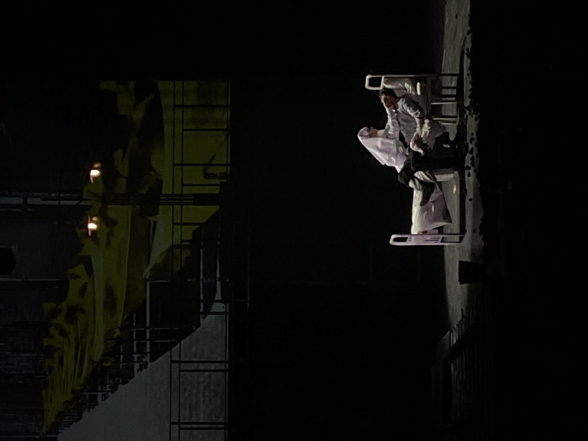 The image shows a dark interior or stage setting, possibly in Заволжье, Russia, with elements suggesting a theatrical production. On the left, there's a dimly lit structure with geometric designs and apparent lighting fixtures. On the right, two individuals are positioned on or near white, minimalist furniture. One figure appears to be wearing a white garment, possibly traditional Middle Eastern attire, while the other seems to be dressed in Western clothing, interacting with the first figure. The overall ambiance is dark and dramatic.