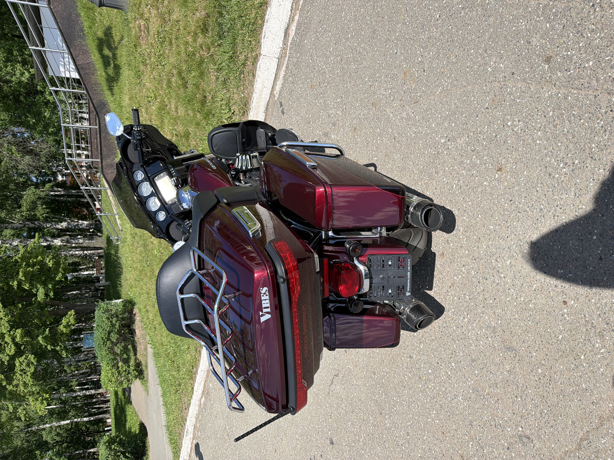 This image depicts a metallic dark red Harley-Davidson touring motorcycle, identified by "HARLEY-DAVIDSON" text on the rear light bar, parked on an asphalt surface under bright sunlight in Votkinsk, Russia. The motorcycle is observed from its rear-right quarter. It features a prominent front fairing equipped with multiple analog gauges and a central digital display, a black passenger backrest, and a chrome luggage rack positioned atop a color-matched rear top case. The top case bears the white lettering "VIBES" and has a short, black antenna extending from its left side. The rear of the motorcycle includes a large red taillight, two smaller black auxiliary lights, and a black license plate frame. This frame contains white Russian text: "ПРЕМИУМ БАЙК" (PREMIUM BIKE) and the contact number "8 800 775 87 78". Dual exhaust pipes with carbon fiber tips are visible at the lower rear.
The environment behind the motorcycle is a park setting. To the left, a paved pathway curves through an area with manicured green bushes and numerous trees, including identifiable white-barked birch trees and other deciduous trees with full green foliage. To the right, a grassy slope rises from a concrete curb bordering the asphalt. A metal railing is visible along the upper portion of this slope, suggesting stairs or a ramp. The clear sky and high sun angle result in sharp shadows cast by the motorcycle and an unseen object or person onto the asphalt. No individuals are present in the image.