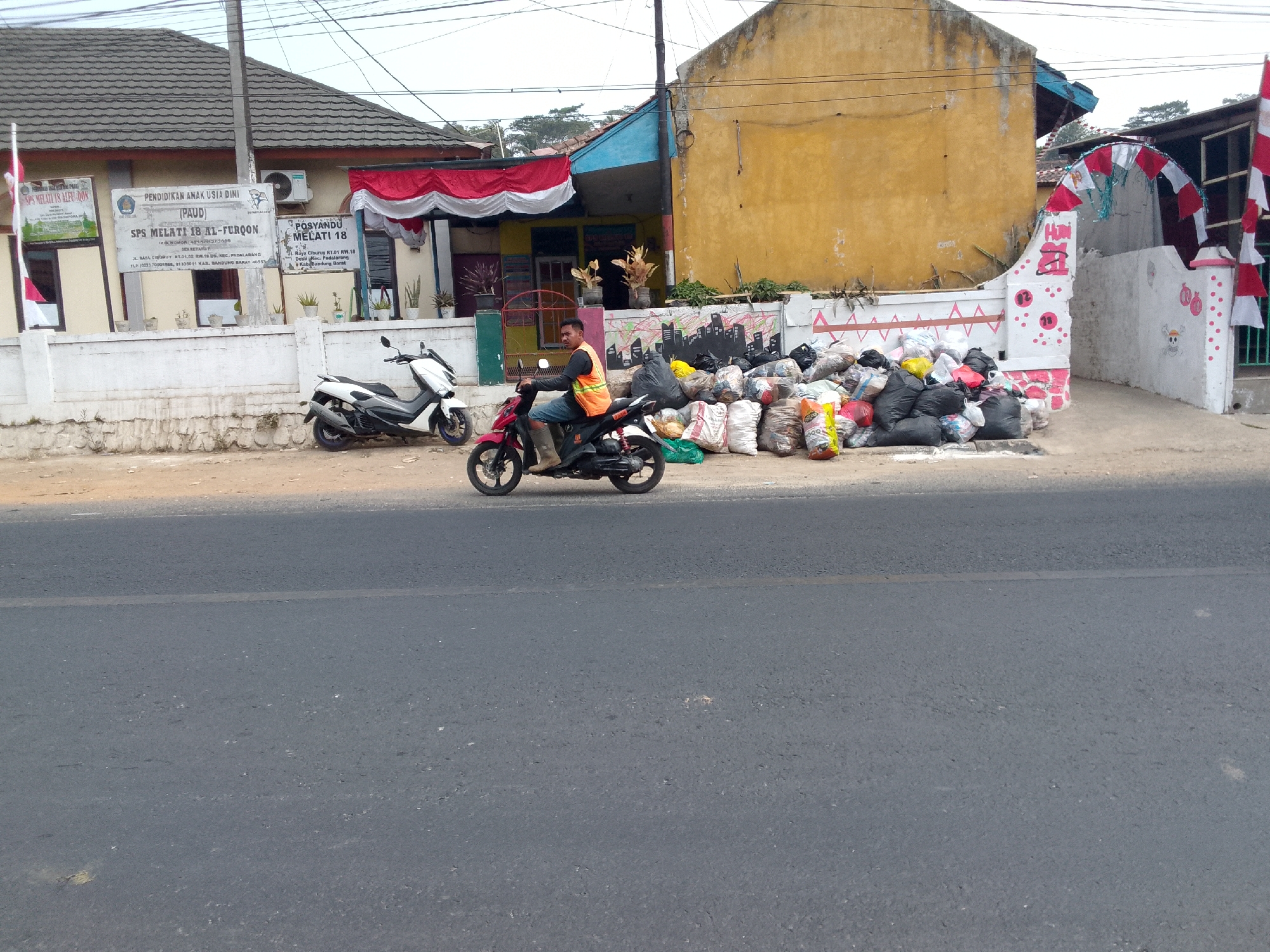 A man wearing an orange safety vest and shorts rides a red motorcycle along a paved road in front of a building with a yellow facade. The motorcycle is positioned on the right side of the road. To the left of the motorcycle, a white scooter is parked. In the background, there is a pile of black and multi-colored garbage bags. The building features a white wall with decorative red and white banners and a mural. A flag on a pole is visible to the left of the building. The scene appears to be in a semi-urban or rural area in Gunungmasigit, Indonesia, given the presence of a school sign and local community center indicators. The road is paved and has a clear lane marking. The environment suggests a sunny day with moderate lighting.