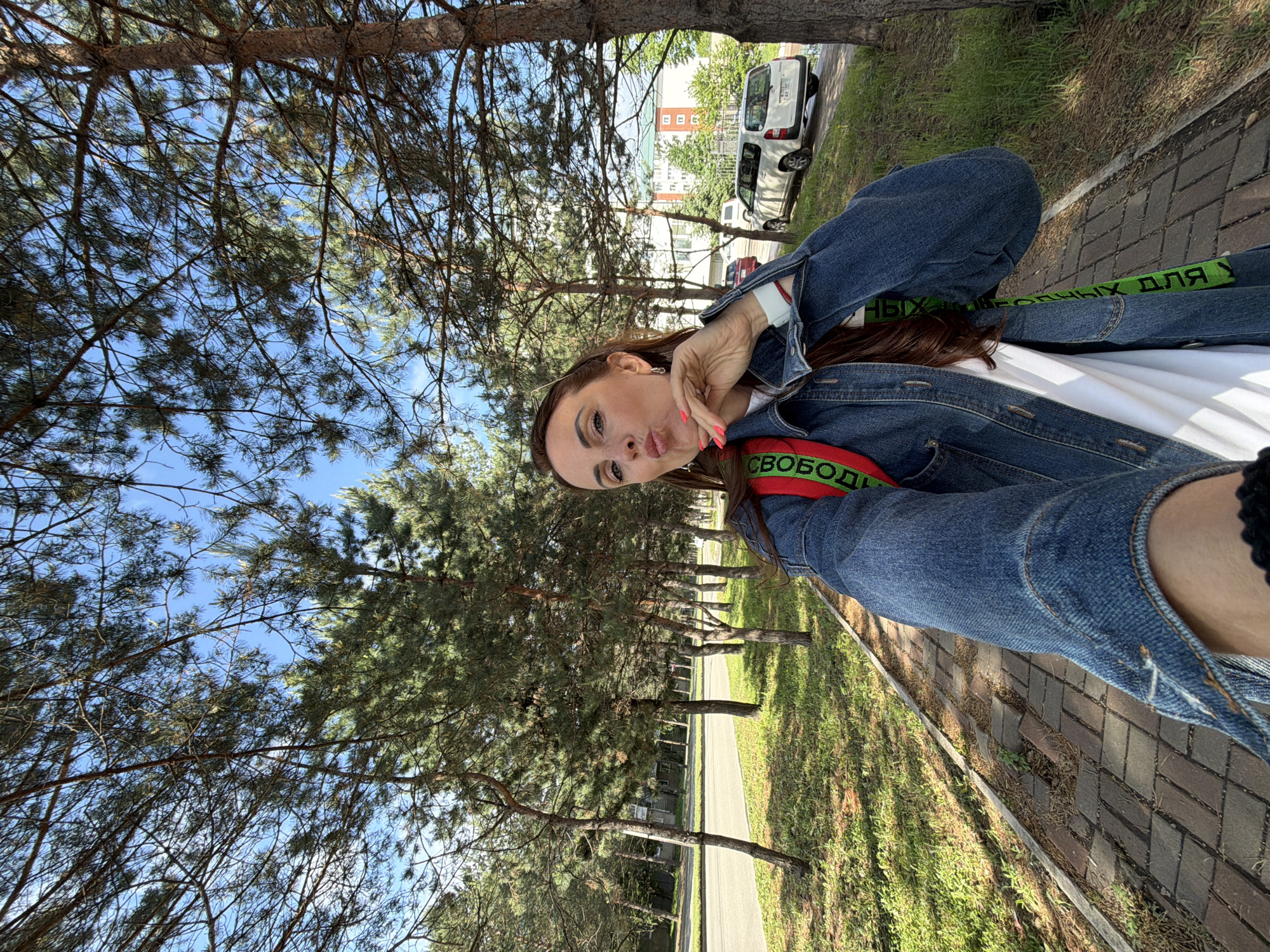 A woman takes a selfie outdoors on a paved walkway lined with trees. She is wearing a denim jacket over a white shirt and a red backpack with green straps. The woman is puckering her lips and gesturing with her hand. In the background, there is a road with parked cars and a multi-story building. The scene appears to be in Birobidzhan, Russia.