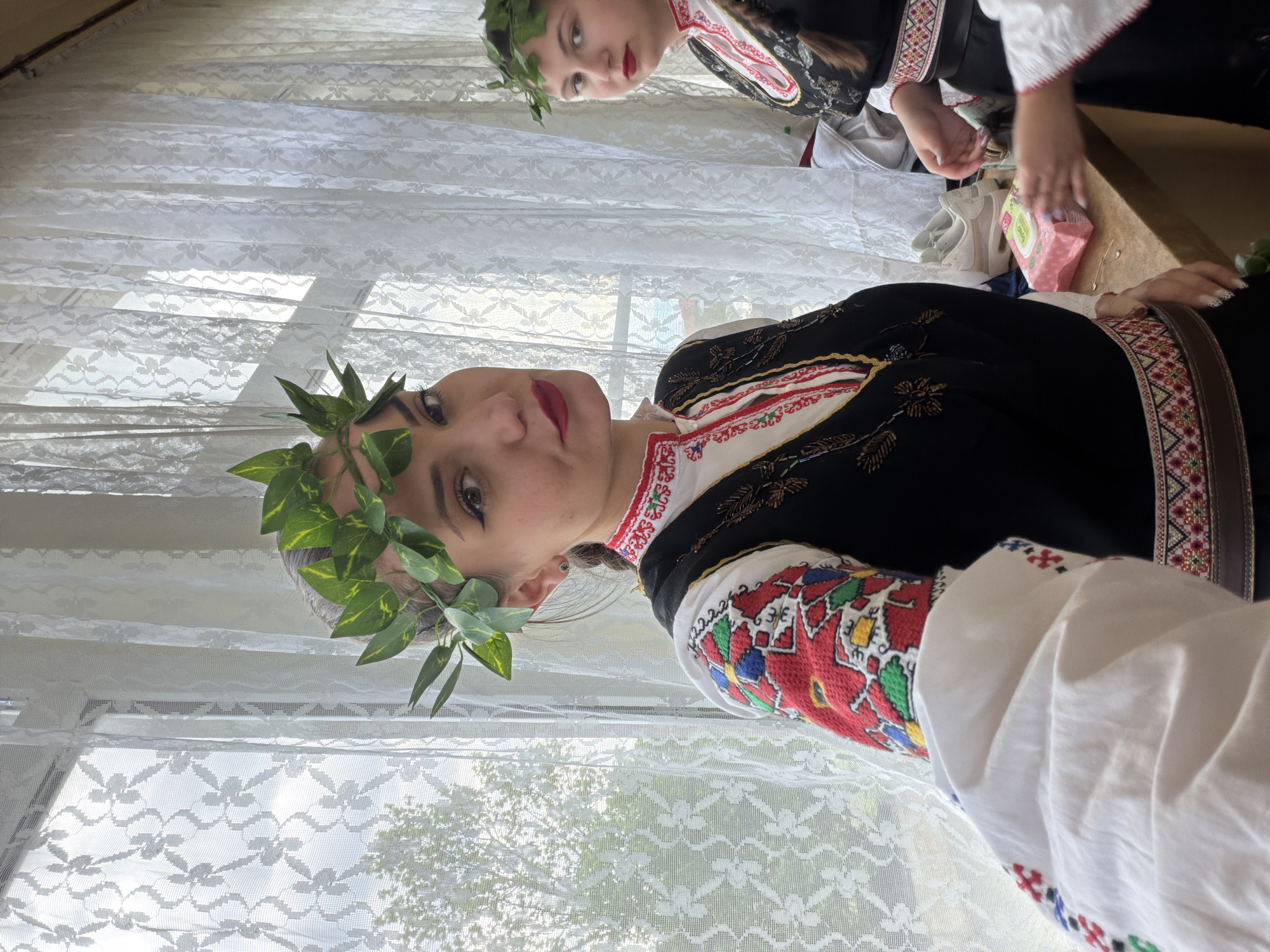 Two women are indoors, wearing traditional Bulgarian clothing. The woman in the foreground is facing the camera with a wreath of green leaves on her head. She is wearing a white shirt with red, yellow, green, and blue embroidered patterns on the sleeves and a black vest with gold designs. The woman in the background is also wearing a wreath and traditional attire. They are near a window covered with white lace curtains. The location is likely in Sofia, Bulgaria, given the traditional clothing.