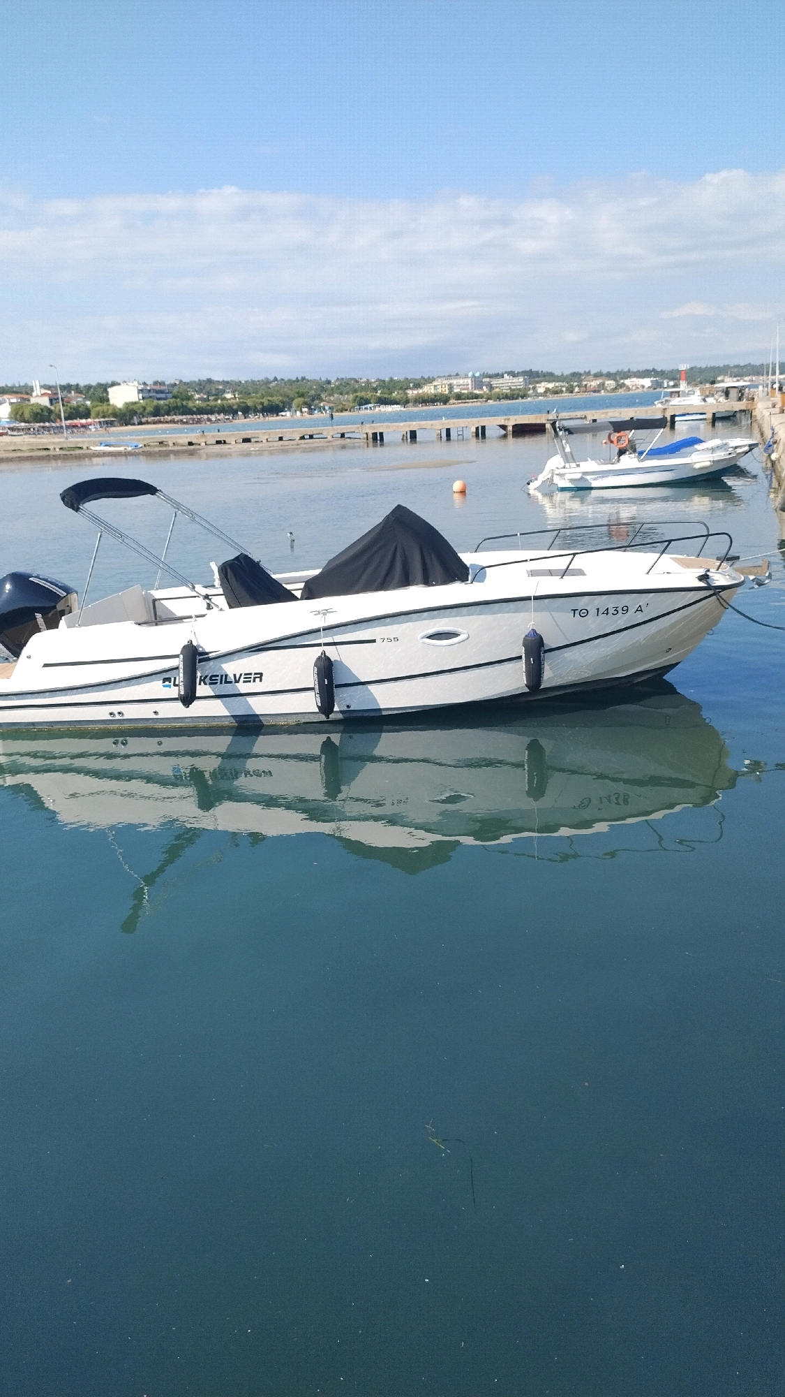 In the city of Neoi Epivates, Greece, a white Quicksilver 755 motorboat is docked in the harbor, its reflection visible in the calm, blue water. The boat, equipped with an outboard motor, has a black canopy and a black cover over a portion of its interior. Two black fenders hang from its hull, and the registration number "TQ 1439 A'" is visible on its side. In the background, a wooden pier extends into the water, with other boats docked and a distant coastline featuring buildings and vegetation under a clear, blue sky with scattered clouds.