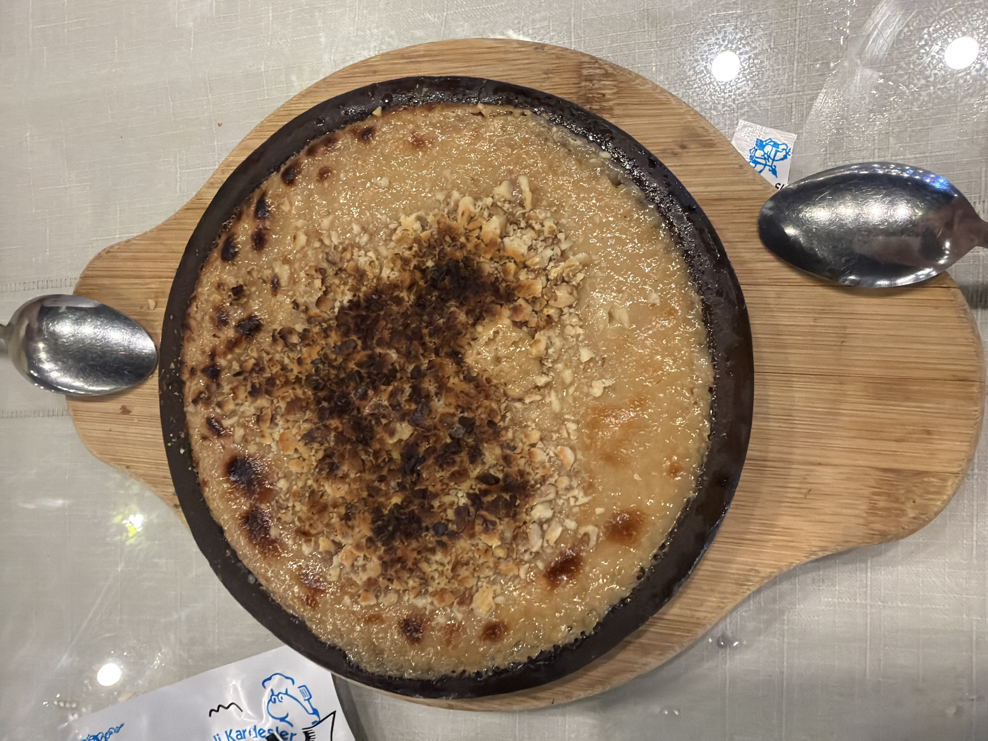 The image shows a close-up, overhead view of a circular, dark-colored dish filled with a creamy, golden-brown dessert topped with chopped nuts and some darker, caramelized bits. The dish is placed on a round, wooden serving board. Two metal spoons are visible, one above and one below the dessert. In the upper left corner, a portion of a white paper packet with blue lettering and an illustration of a person is present. A white textured tablecloth is underneath the serving board. The overall impression is of a dessert served in a restaurant or home dining setting. There are no individuals or discernible city landmarks visible to indicate a specific location in Kocaeli, Turkey.