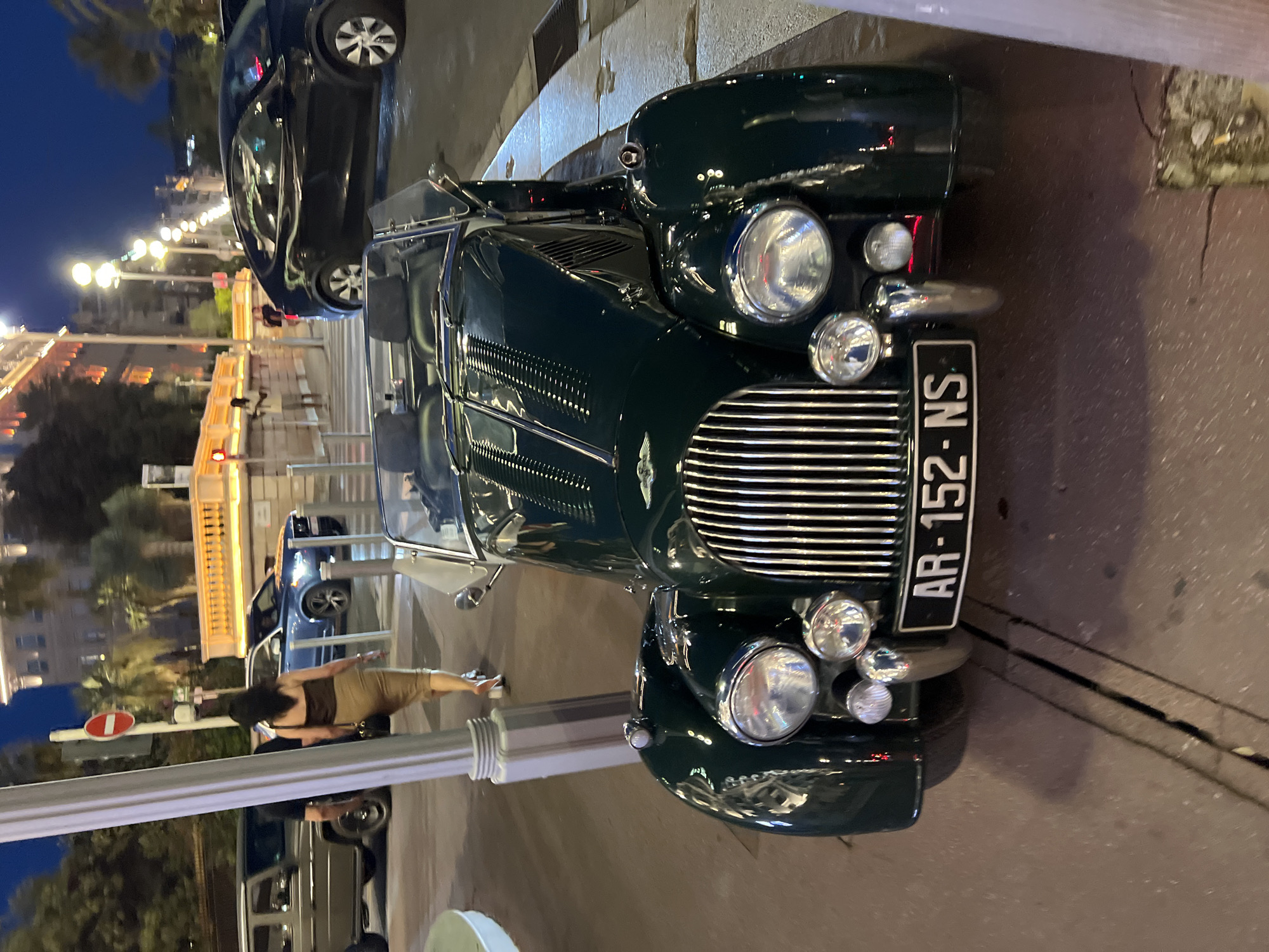 An evening scene on the Promenade des Anglais in Nice, France, captures a dark green vintage convertible automobile, possibly a Morgan, parked on the sidewalk. The car features a prominent chrome grille, dual headlights, and two smaller auxiliary lights. Its license plate reads "AR-152-NS". Behind the car, a woman in a light brown skirt and dark top walks away from the camera, carrying a black purse. In the background, other vehicles are visible, including a grey SUV and a dark sedan. The cityscape is illuminated by streetlights, with the architecture of Nice's beachfront buildings visible. The time appears to be dusk or early evening, indicated by the lit streetlights and the ambient light.