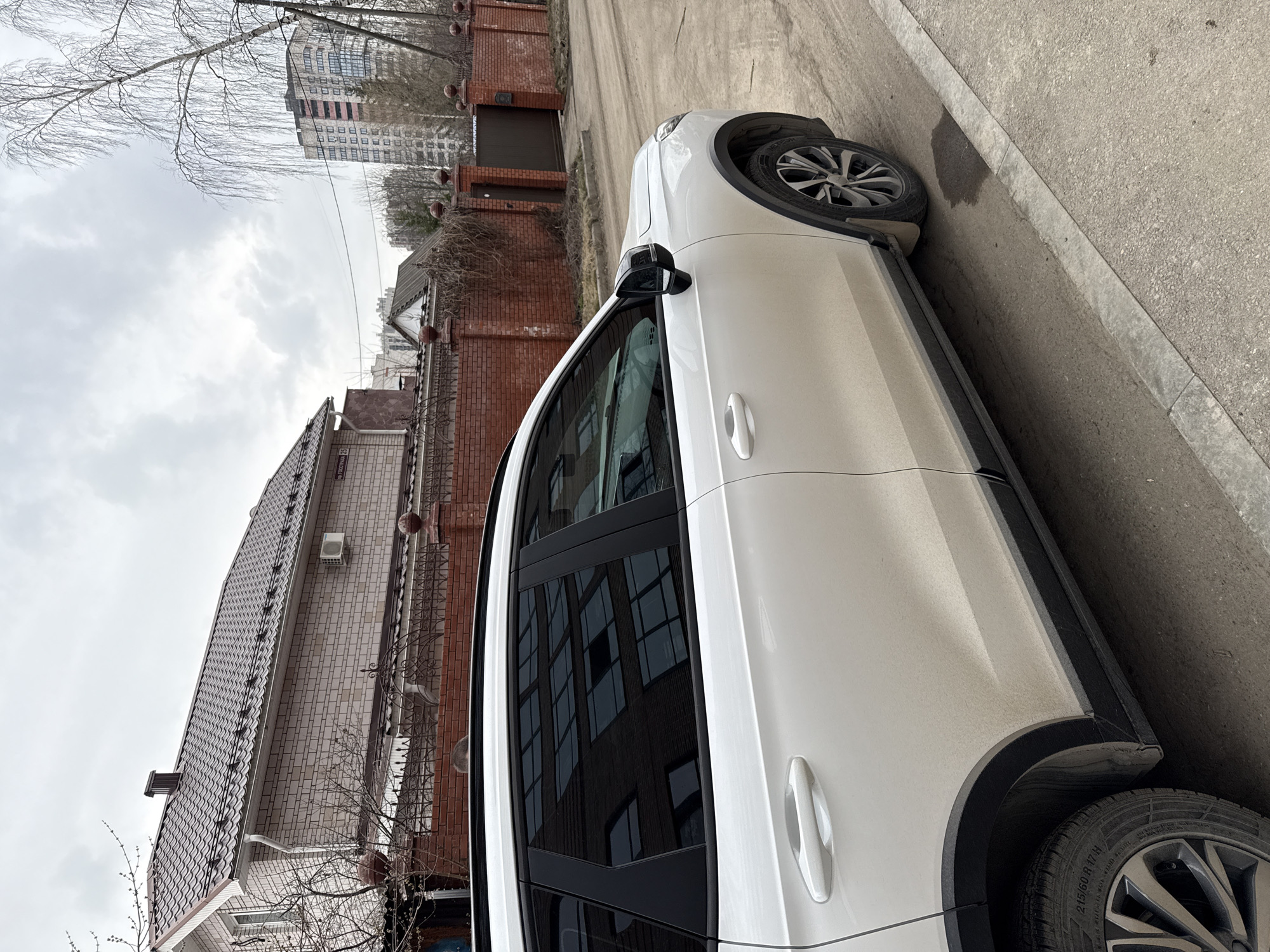 A white car is parked on the side of a street in Izhevsk, Russia. The background shows a residential area with brick houses and apartment buildings under a cloudy sky. The street is paved and appears slightly dirty. The focus is on the car and the surrounding urban environment.