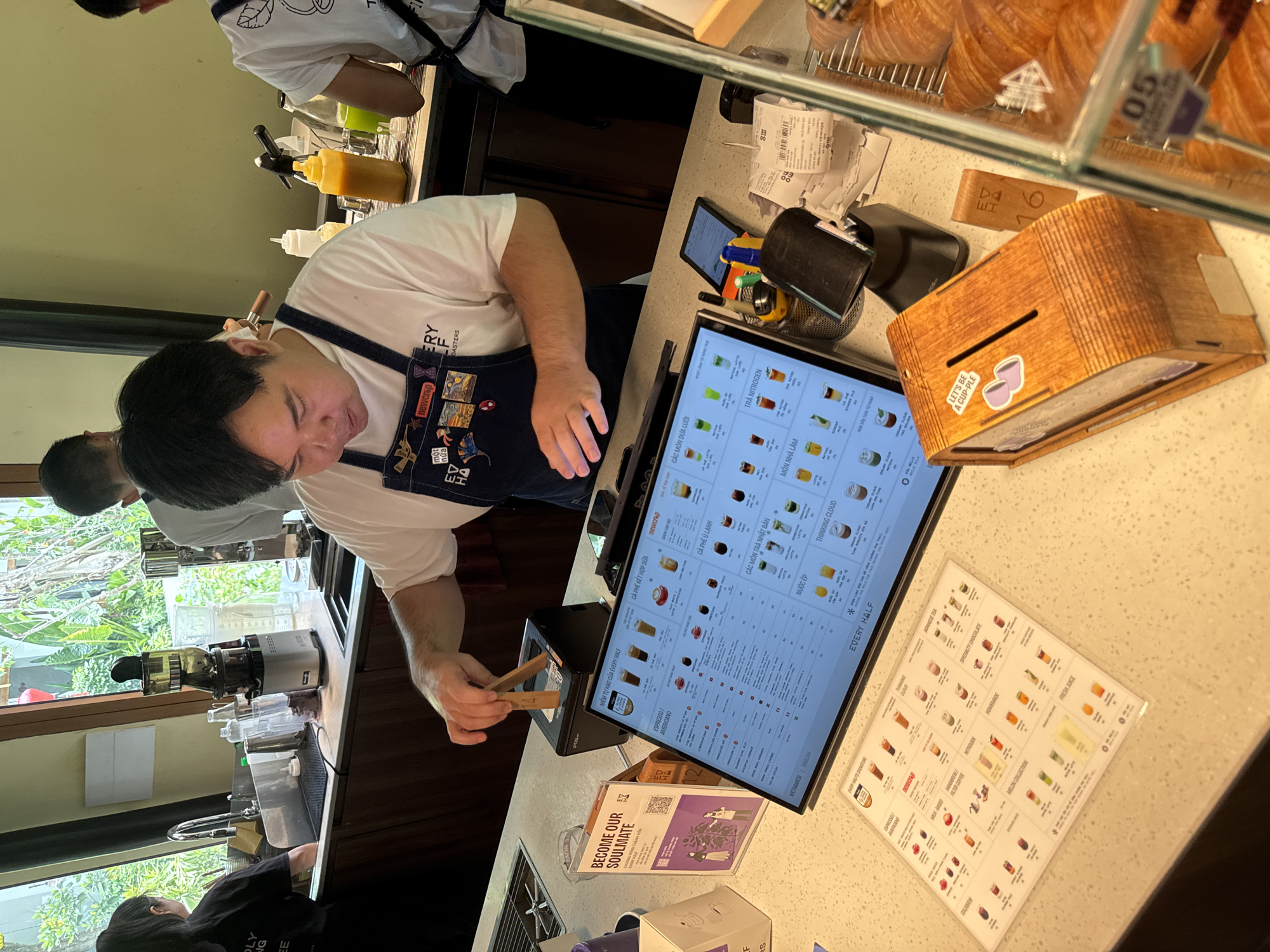 The image shows a coffee shop in Hoi An, Vietnam, with a barista operating a touchscreen menu and cash register. The barista, a man with dark hair wearing a white t-shirt and a dark apron, is in the process of selecting items from the menu, indicated by his hand holding wooden tongs near the register. The menu displays a variety of drinks, including espresso, americano, milk-based coffees, Japanese teas, and specialty chocolates, with prices in Vietnamese Dong. A second barista, partially visible in the background wearing a white t-shirt and a dark apron, is attending to customers. The coffee shop appears modern and well-equipped, with a clean and organized counter area. There is a wooden donation box on the counter that reads "LET'S BE A CUP-PLE". Croissants are visible in a display case to the right. The overall atmosphere suggests a busy, efficient establishment catering to coffee and pastry lovers.