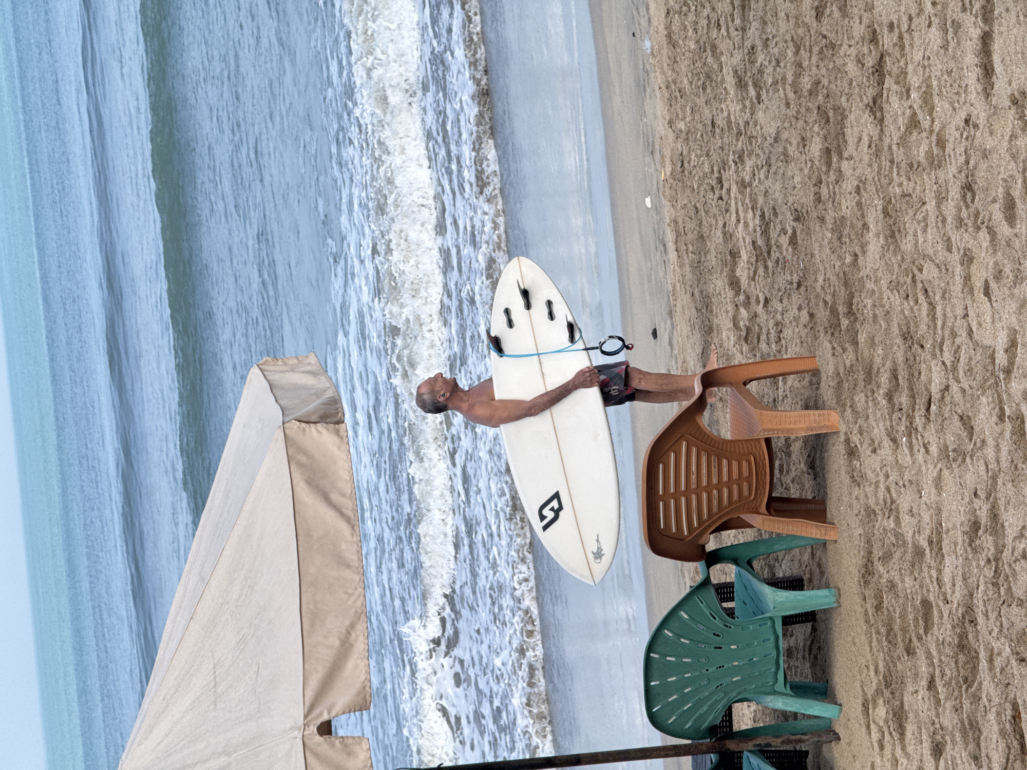 An elderly Caucasian man stands on a sandy beach in Kuta, Indonesia, holding a white surfboard. He is wearing patterned swimming shorts and appears to be barefoot. The man is positioned under a large, tan beach umbrella, and his body is partially obscured by the umbrella's pole. The surfboard is white with black fins and a blue leash. In the foreground, two plastic chairs, one green and one orange, are partially buried in the sand. The ocean is visible in the background, with moderate waves and white foam. The sky is a pale, overcast blue.